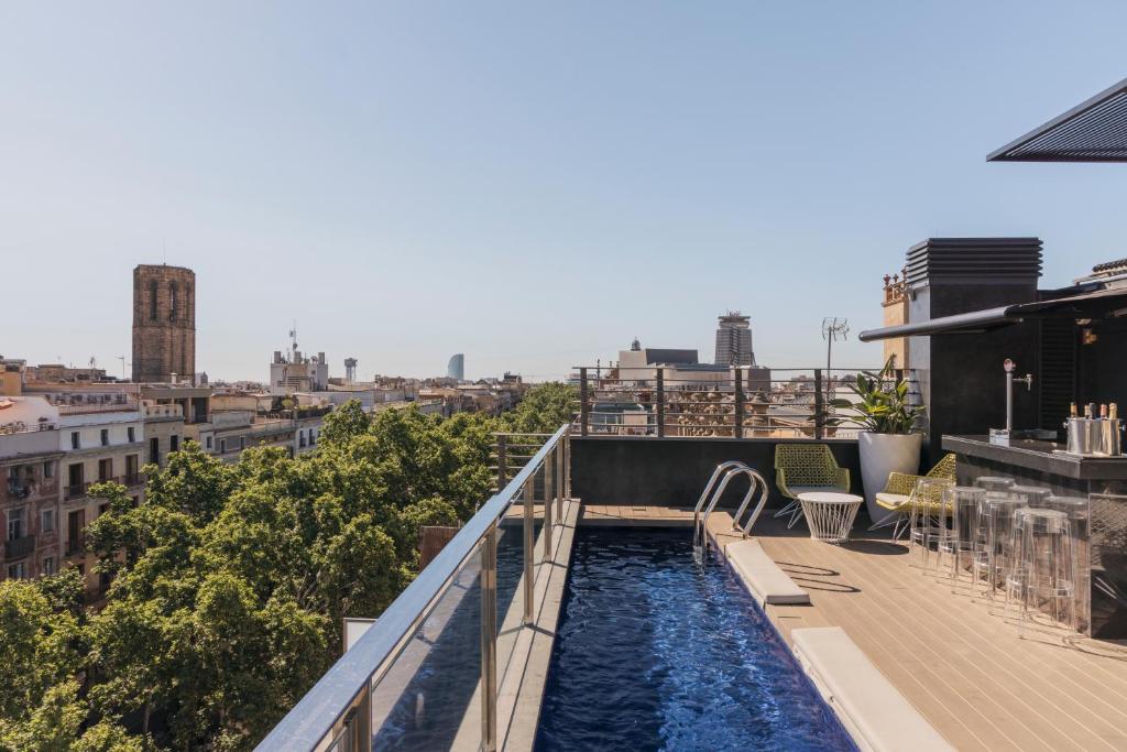 Se ve una terraza con una piscina alargada y tumbonas, rodeada de edificios y árboles al fondo.