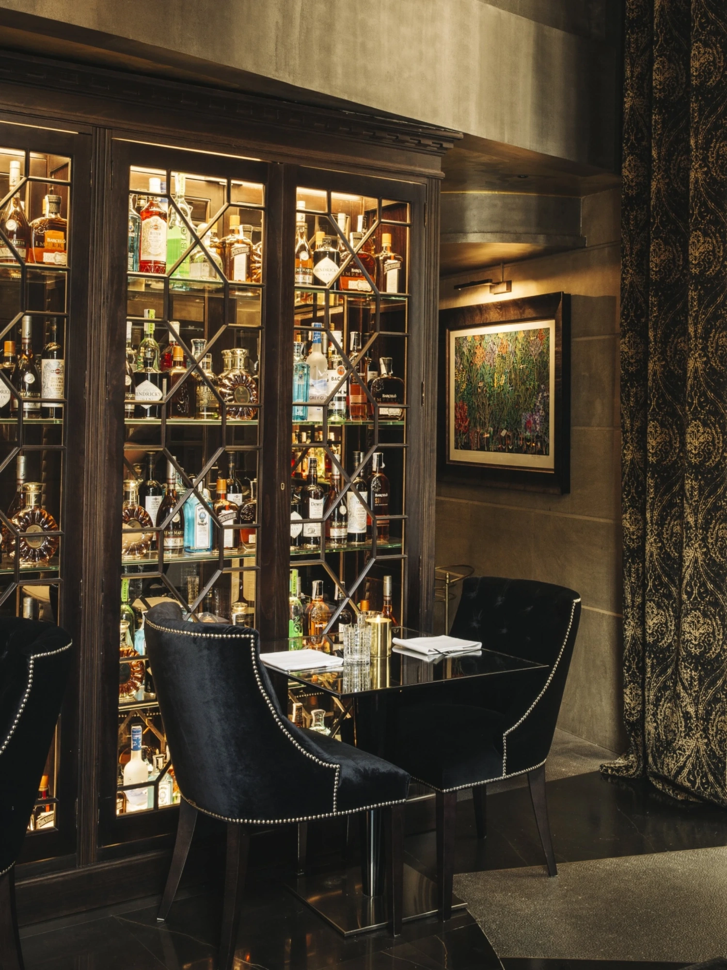 Un bar interior muestra una vitrina con botellas y mesas con sillas cerca del muro decorativo.