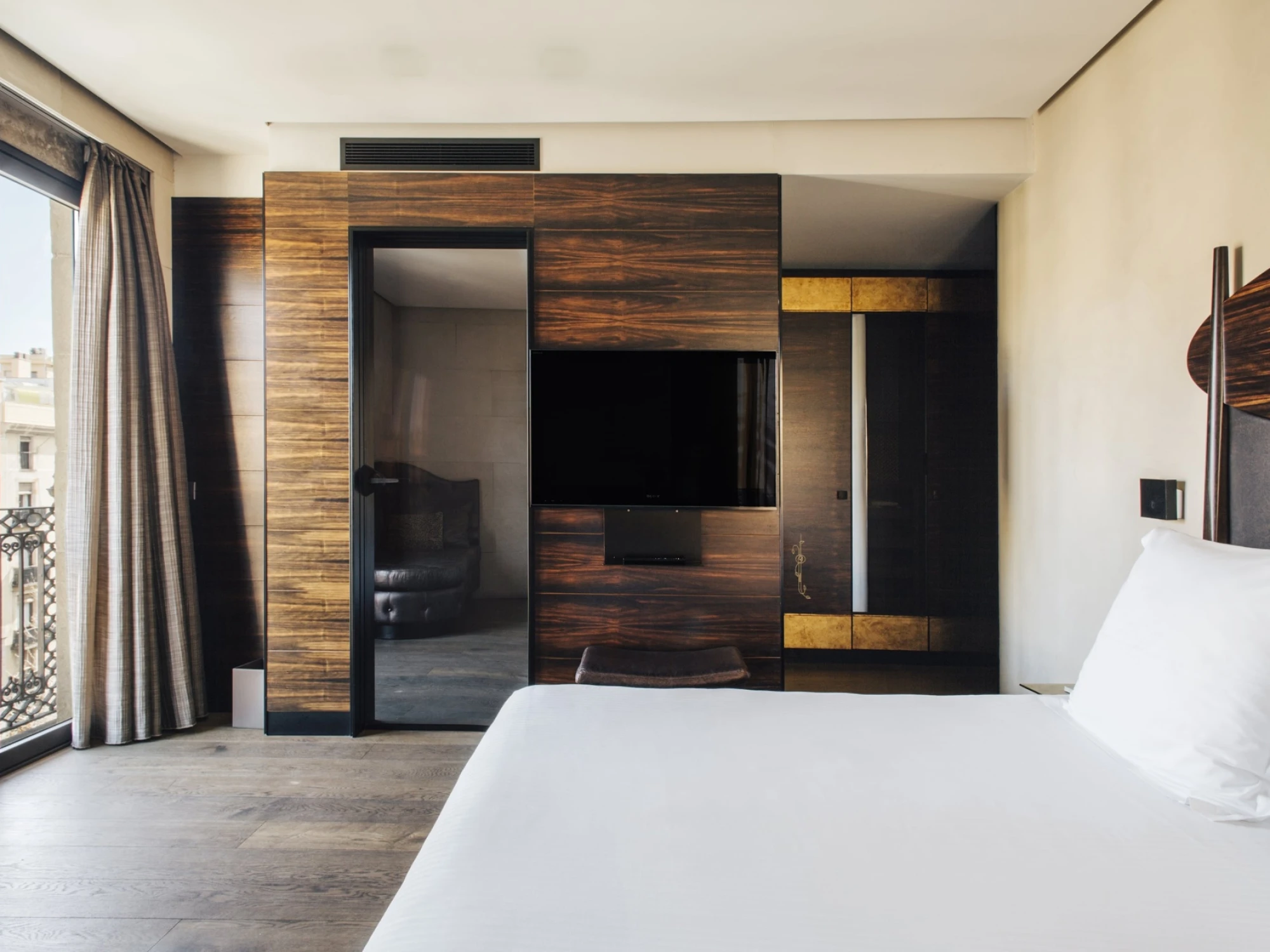 Se ve una cama y un televisor empotrado sobre un panel de madera, con cortinas y una puerta de cristal cerca.