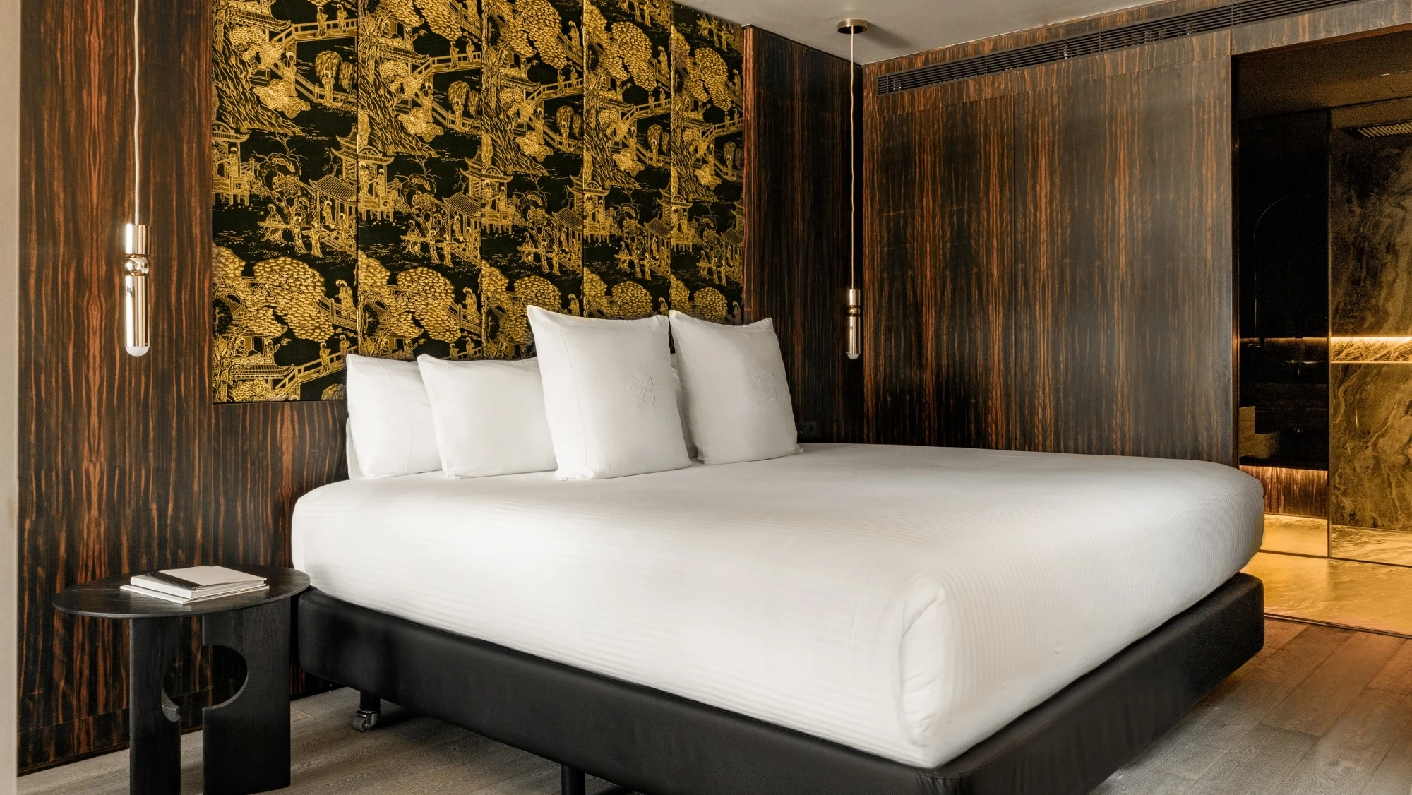 Se ve una habitación con una cama doble con ropa de cama blanca, mesitas laterales oscuras y una pared decorativa detrás de la cabecera.