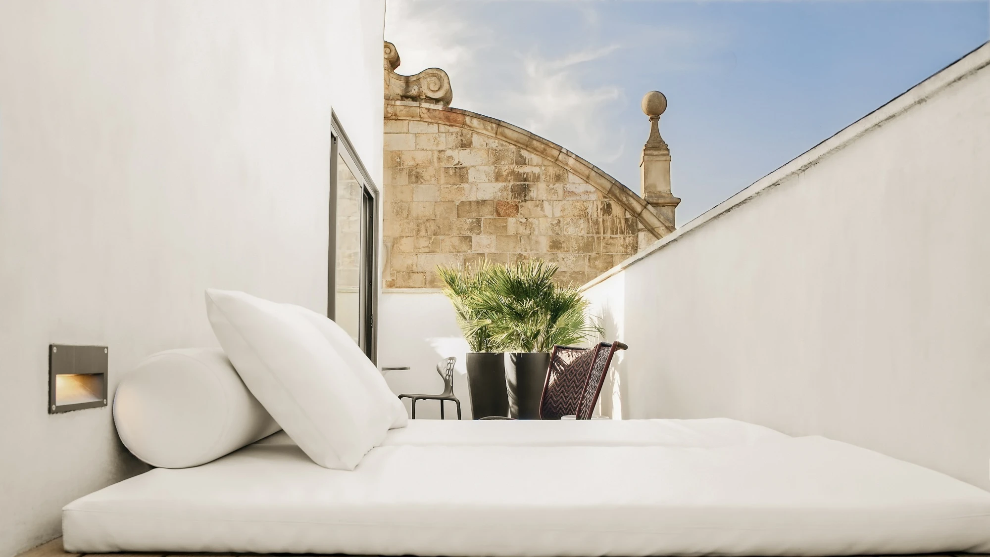 Se ve una cama con ropa de cama clara sobre una terraza con paredes blancas y plantas en macetas.