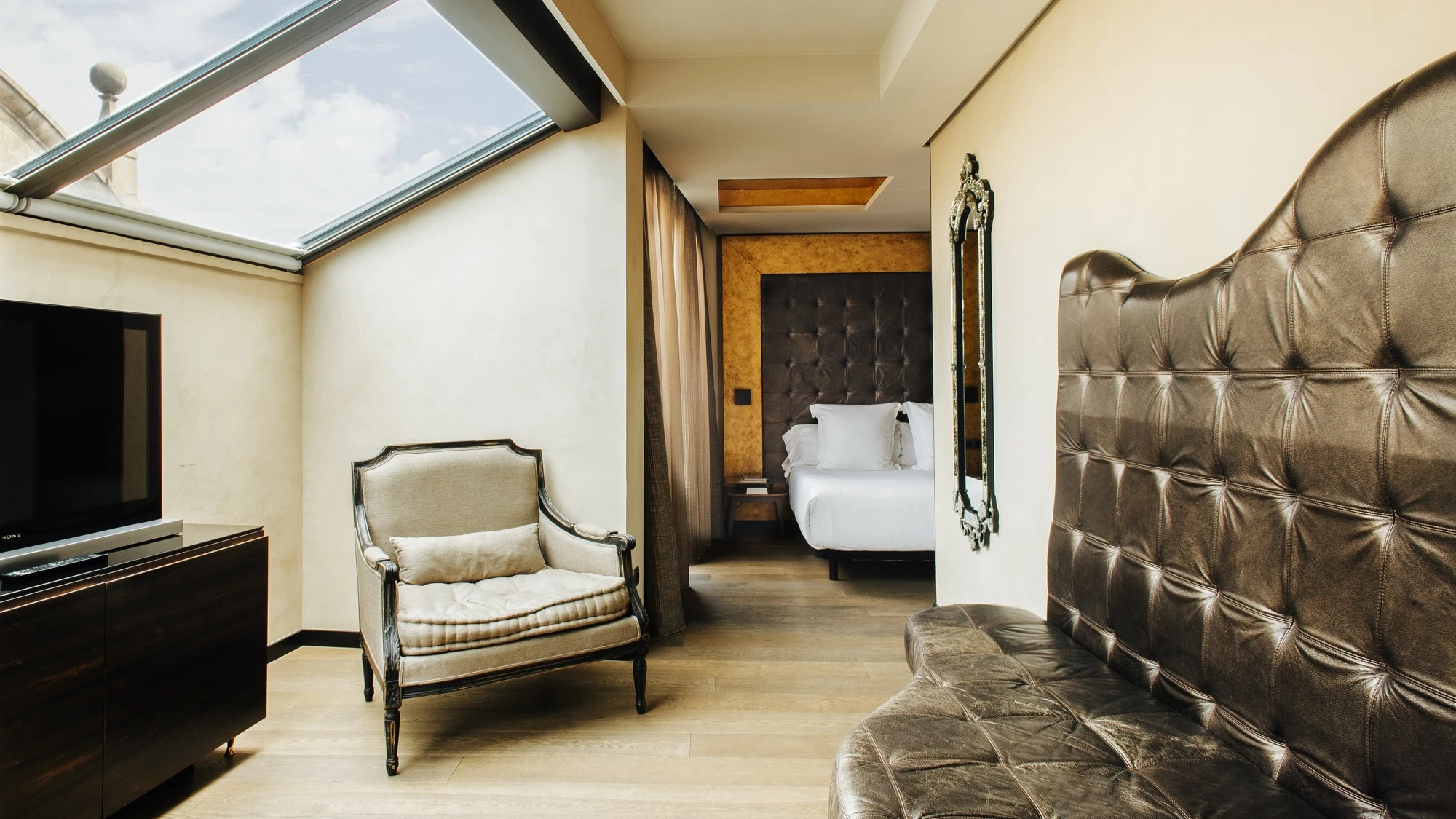 La imagen muestra una sala tipo suite con cama, un sillón junto a una ventana y un sofá tapizado con cojines decorativos.
