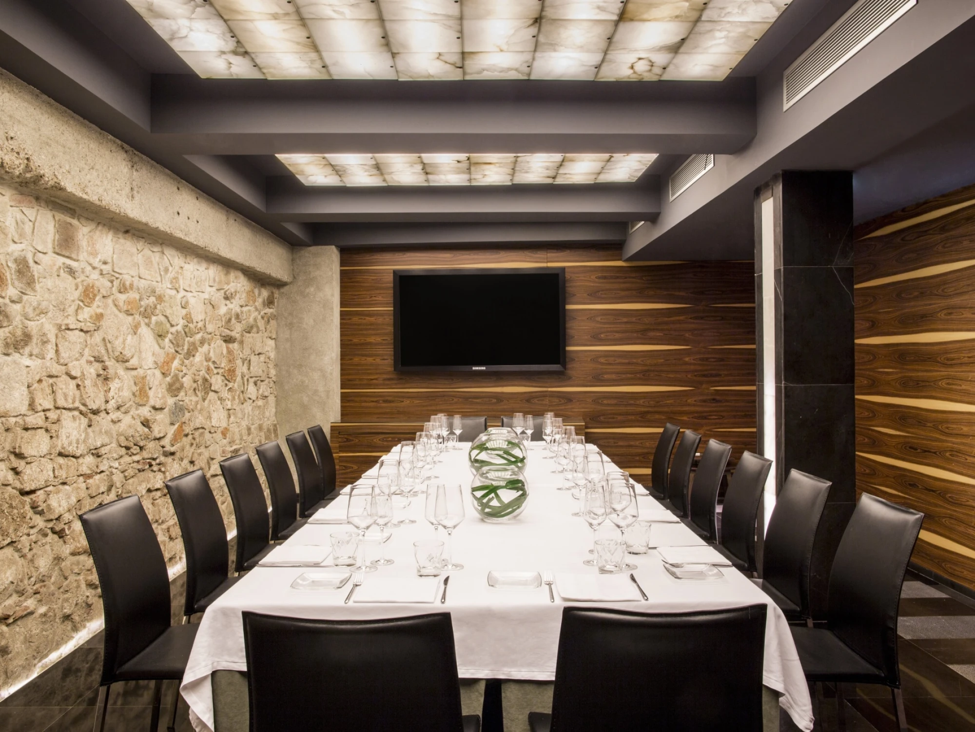 Se ve una sala de reuniones con una mesa larga cubierta y sillas alrededor, con una pared de piedra y un televisor montado.