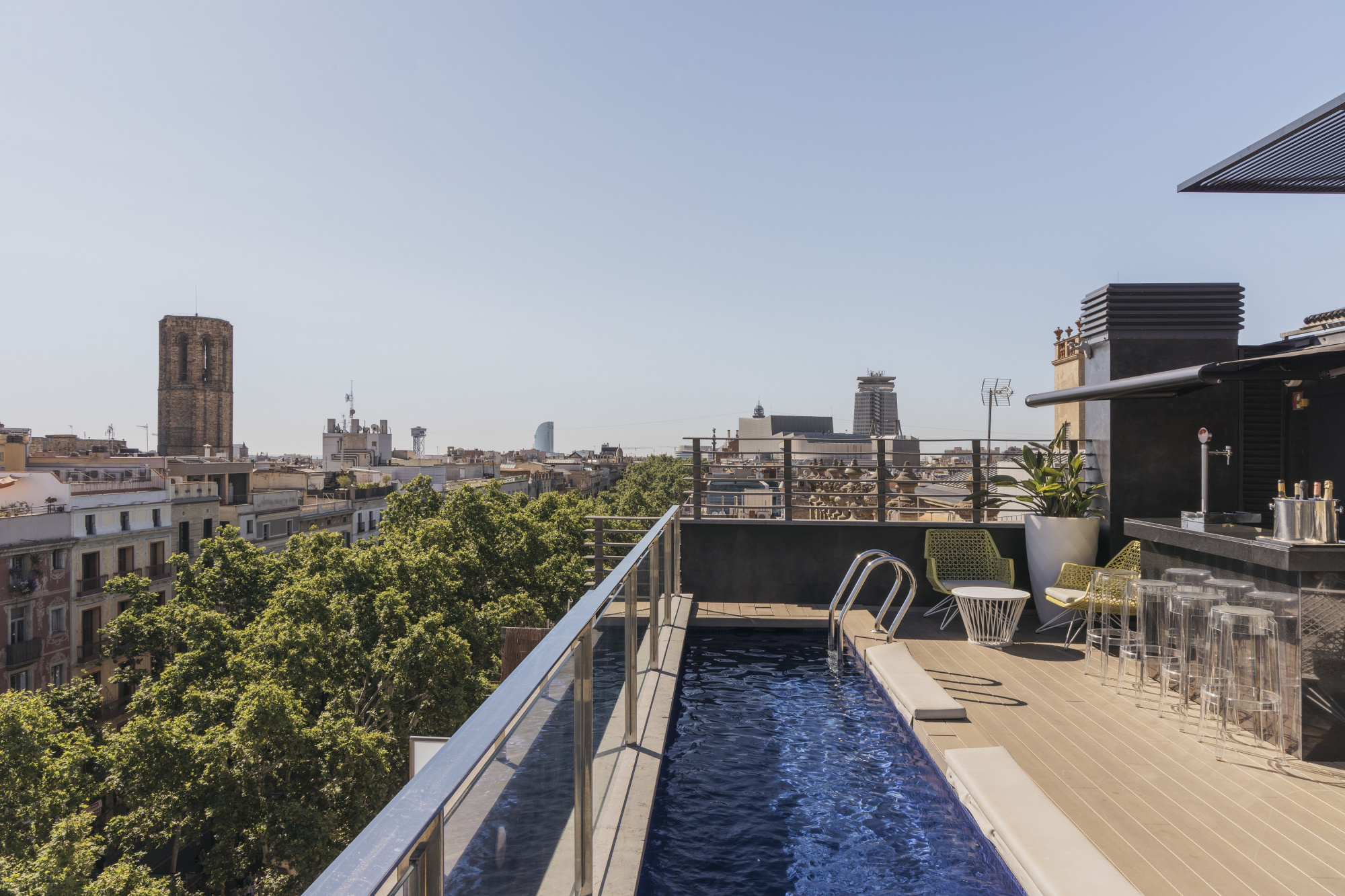 Se ve una piscina exterior estrecha en una terraza con tumbonas y barandilla, con el skyline urbano al fondo.