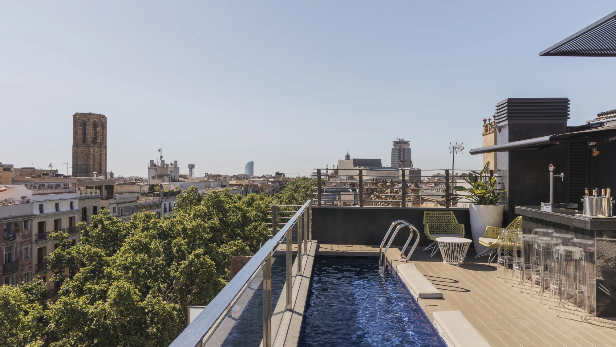 Piscina exterior en una terraza con tumbonas y vista urbana al fondo.
