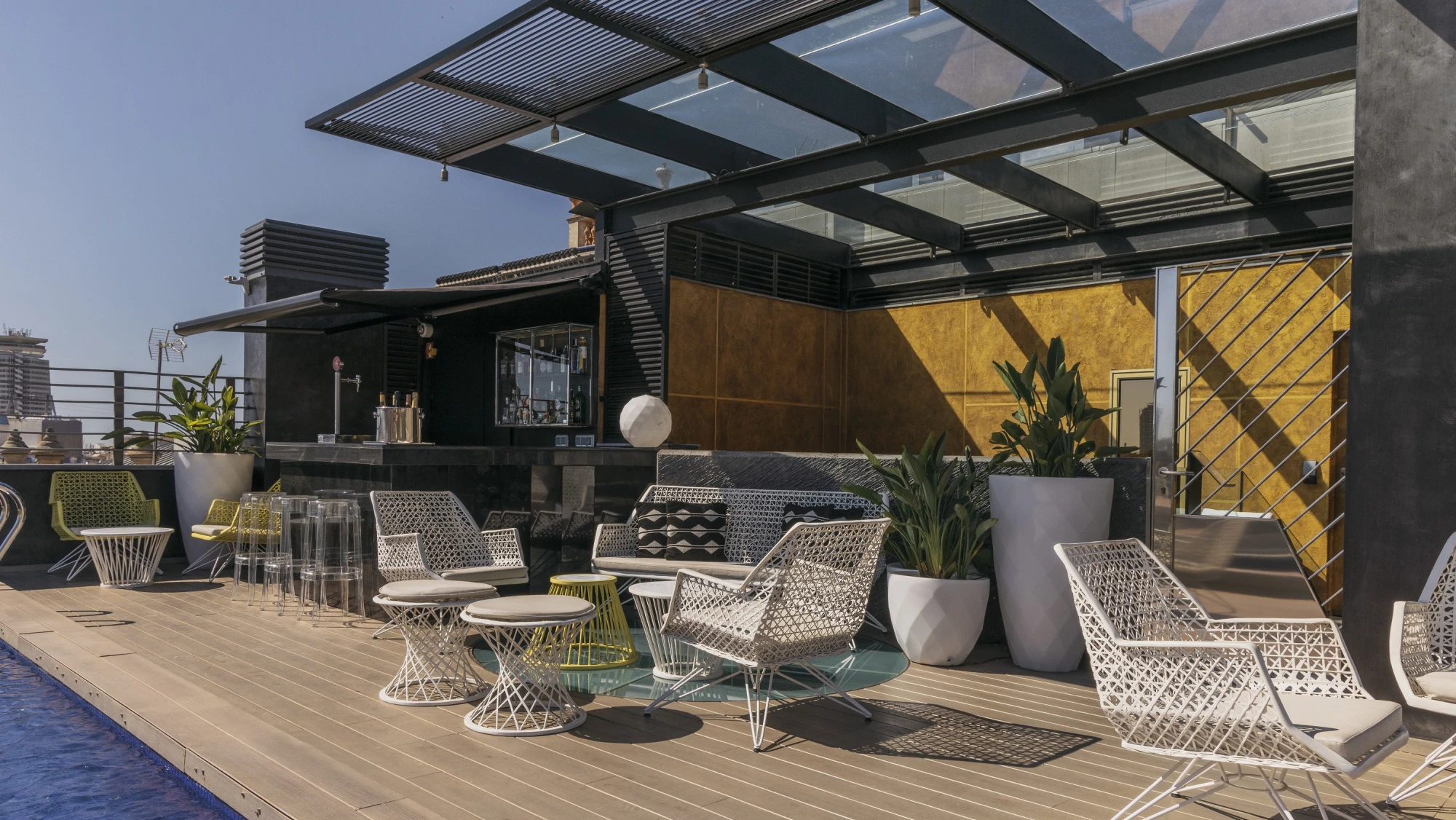 La imagen muestra una terraza con sillas y mesas de malla metálica y plantas en macetas sobre el pavimento de madera.