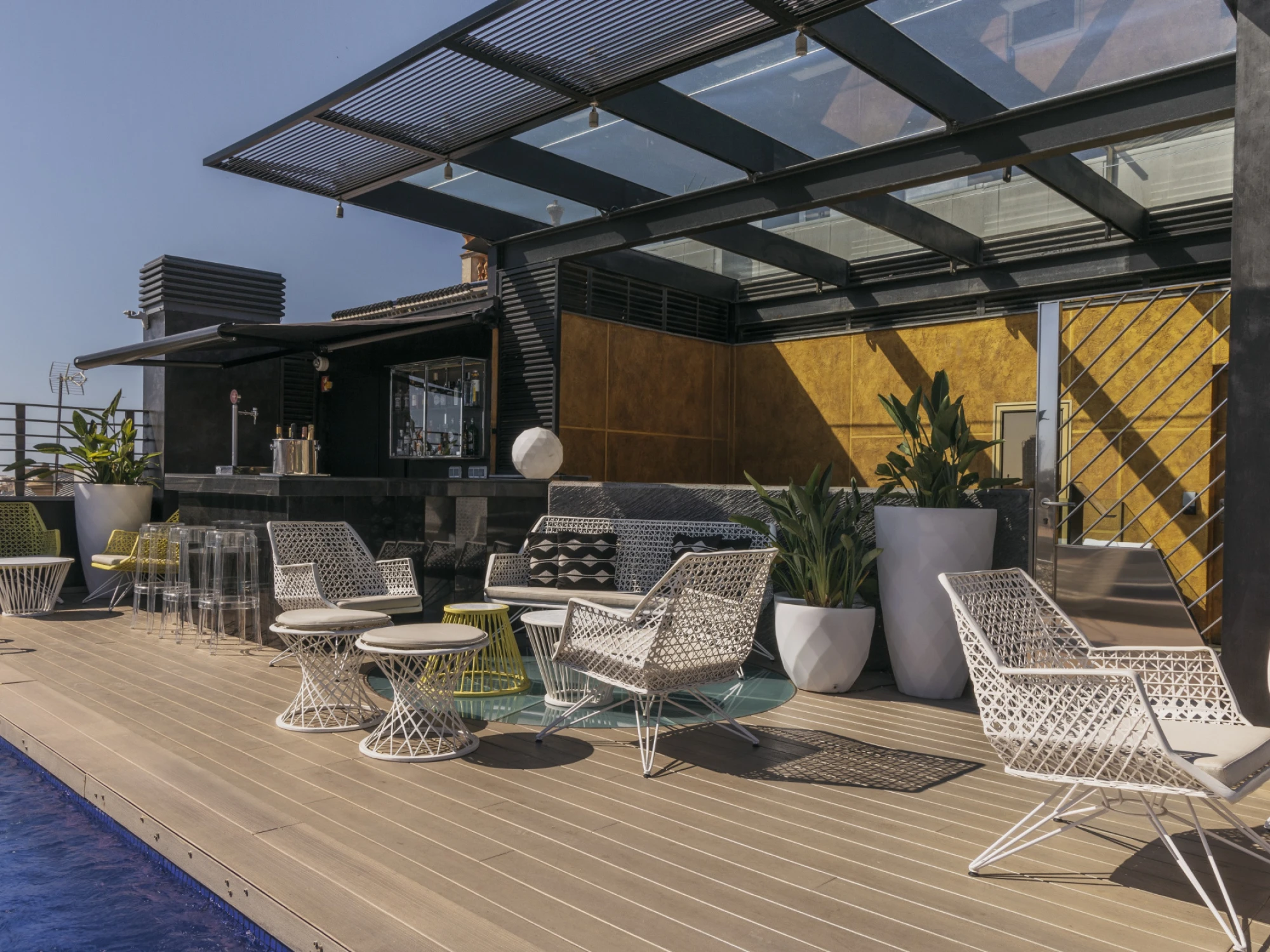 Se ve una terraza exterior con tumbonas, mesas pequeñas y plantas junto a una piscina al aire libre.