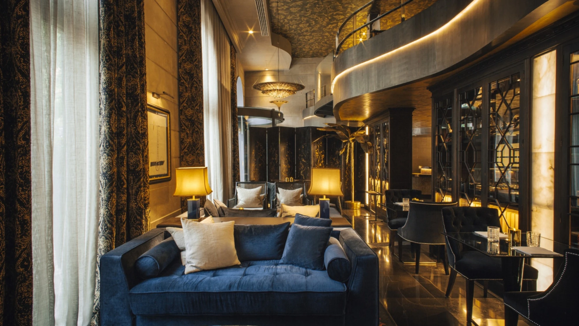 Se ve un salón elegante con sofá azul, cojines y lámparas de pie junto a una pared decorativa.