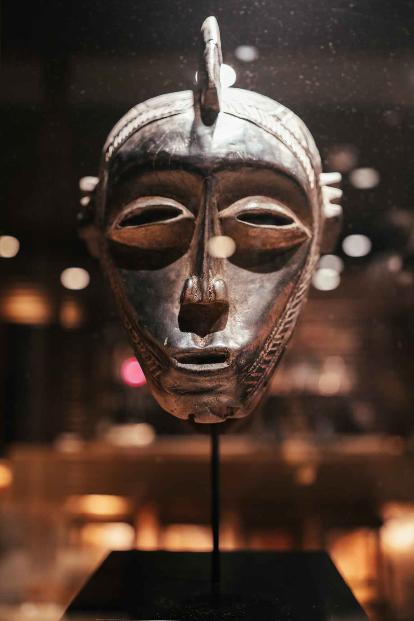 Se ve una máscara africana de metal sobre un soporte, con luces desenfocadas al fondo.