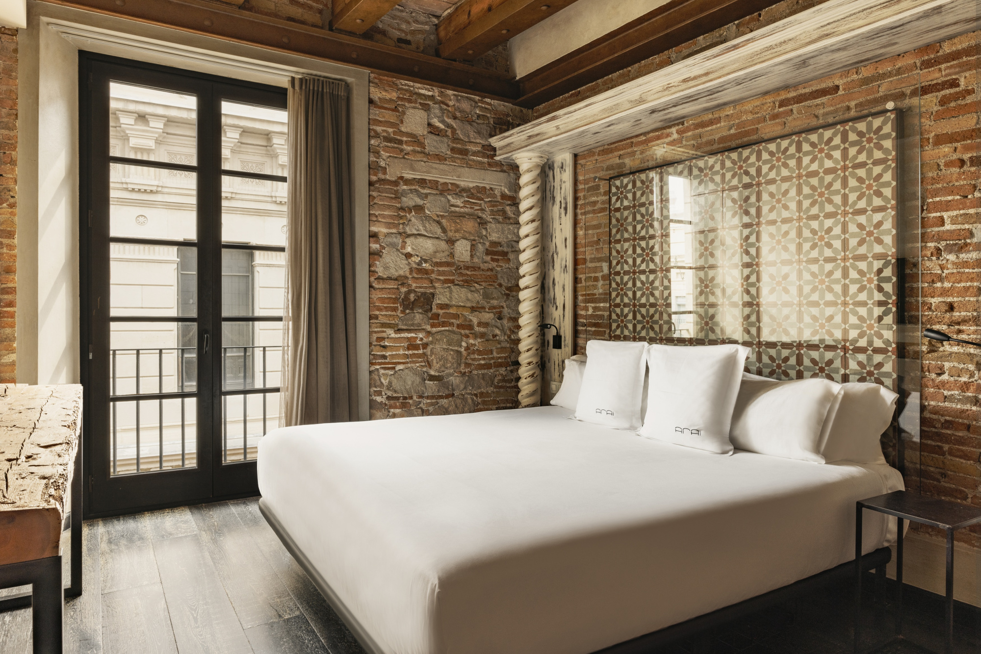 Se ve una cama con ropa de cama blanca y una pared de ladrillo vista junto a una puerta con cortinas.