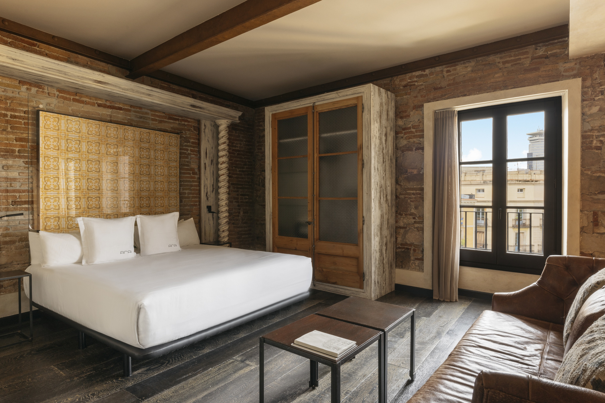 Se ve un dormitorio con una cama doble, un sillón, una mesa auxiliar y ventanas con luz natural.