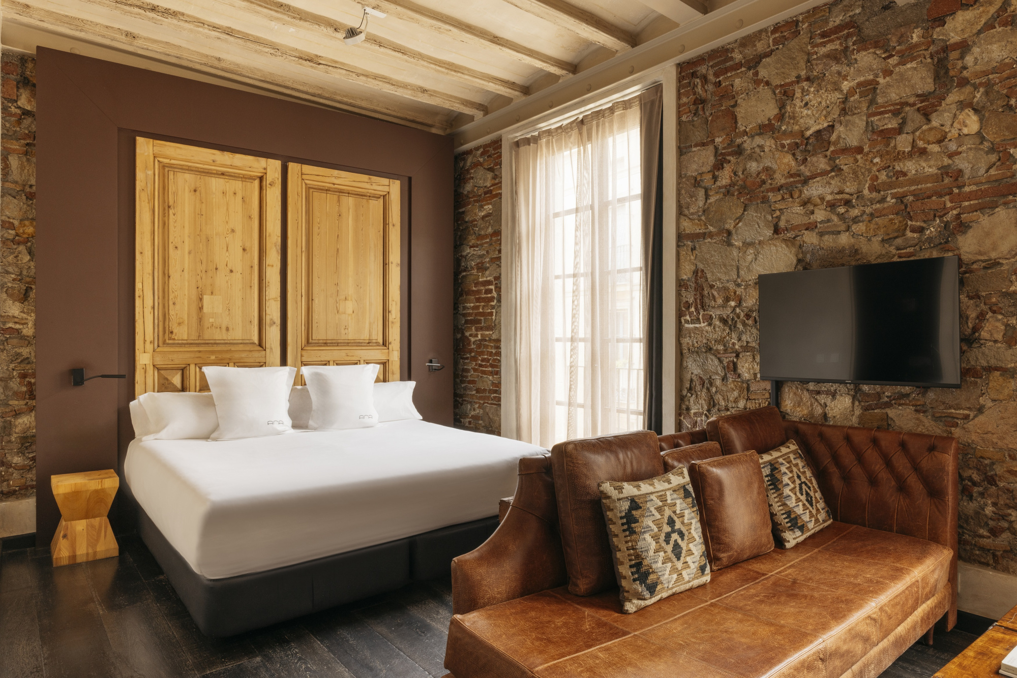 Se ve una cama doble con ropa de cama blanca, un sillón de cuero y una pared de piedra con una televisión.