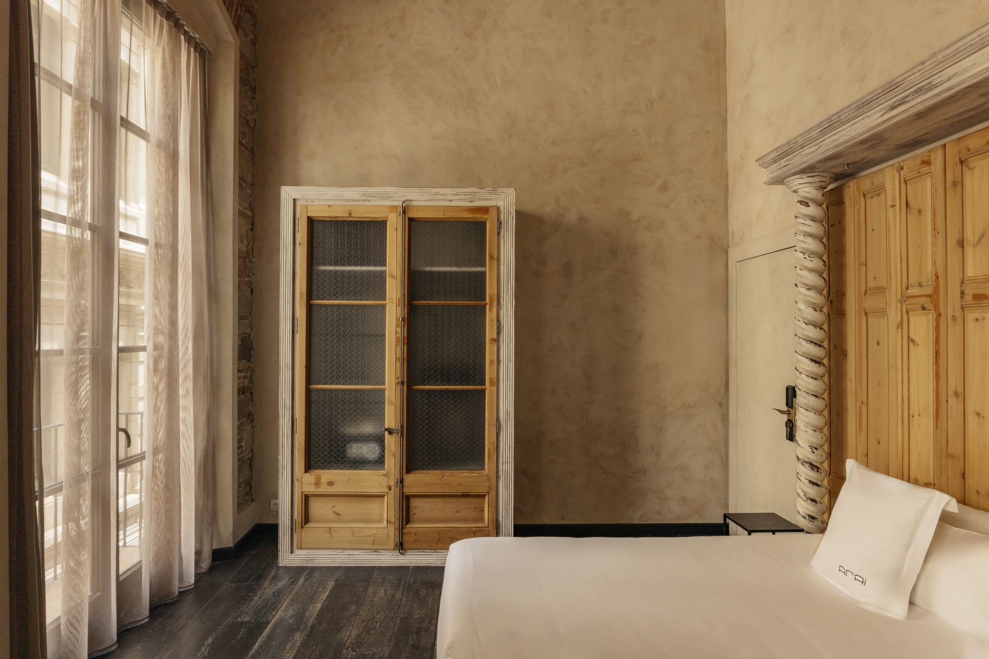 Se ve una cama con almohadas, un armario de madera y una ventana con cortinas en una habitación luminosa.