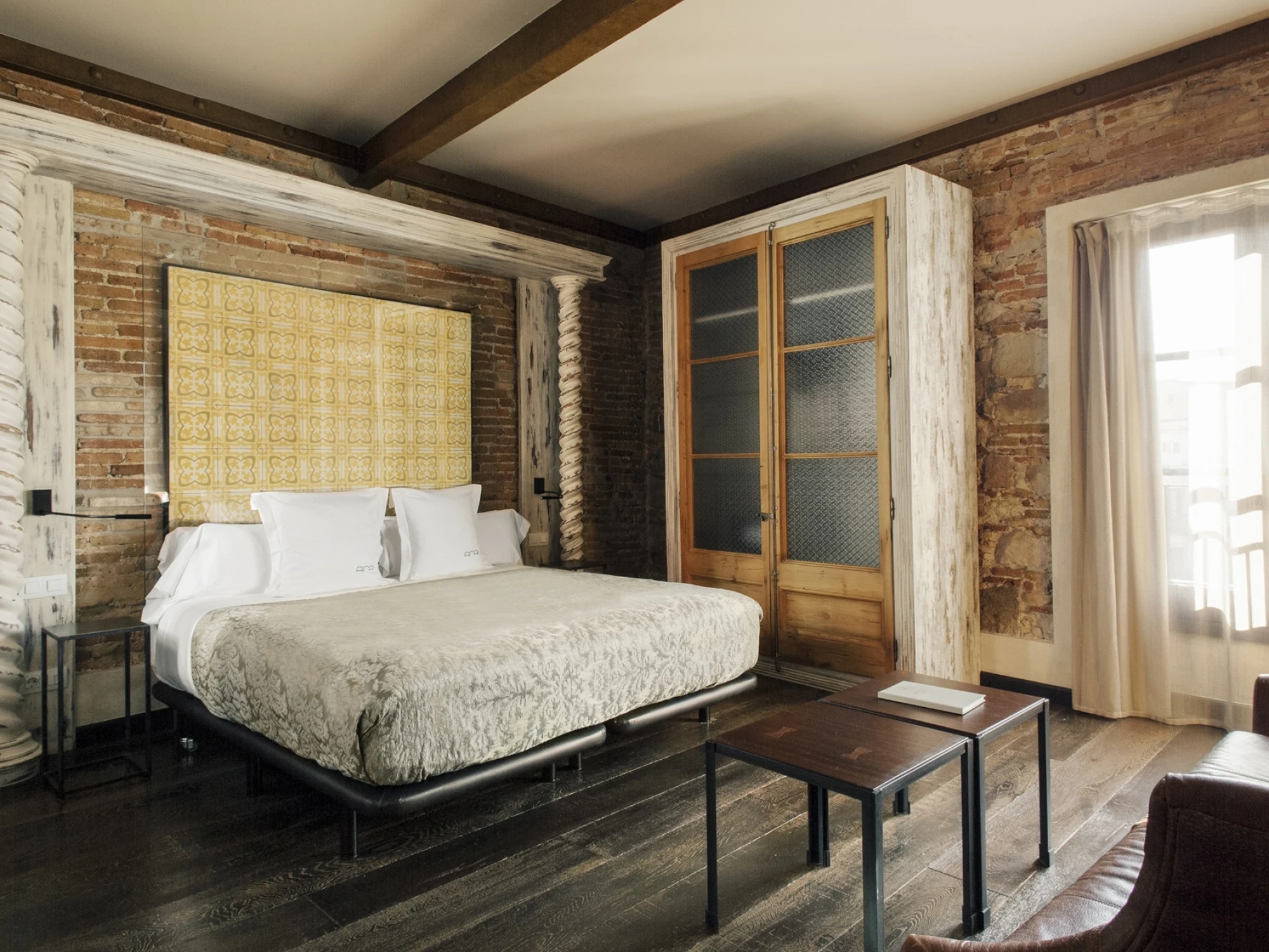 Se ve una cama doble con mesitas y una pared de ladrillo junto a una ventana con cortinas.