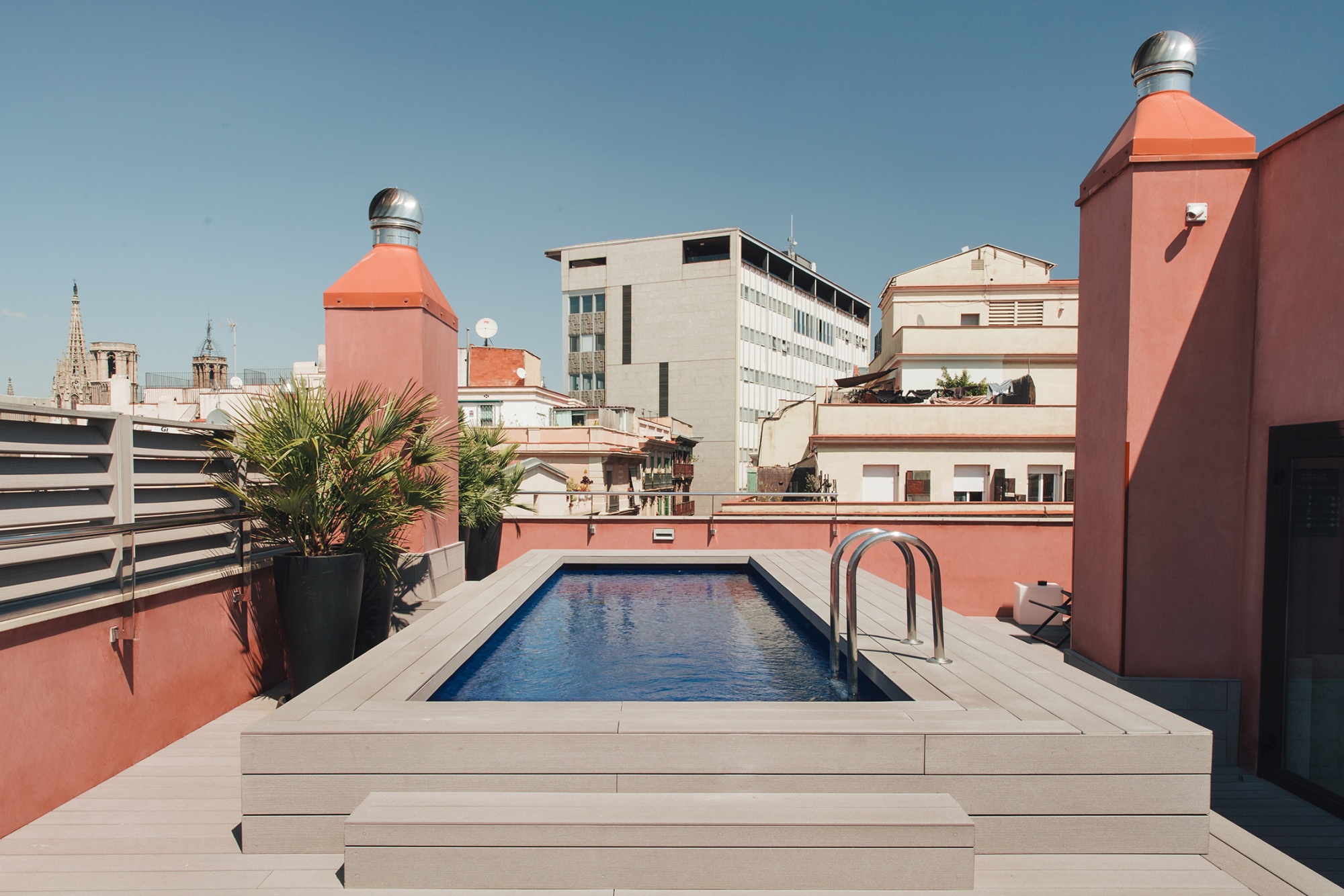 Se ve una piscina rectangular en una terraza con paredes de color rojo y edificios al fondo.