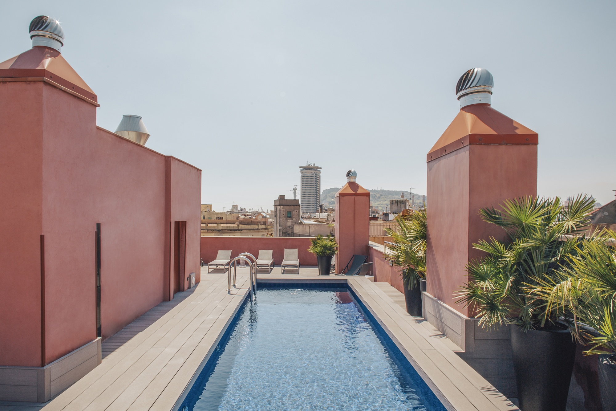 Se ve una piscina rectangular en una terraza con tumbonas y plantas junto a paredes de color rojo.