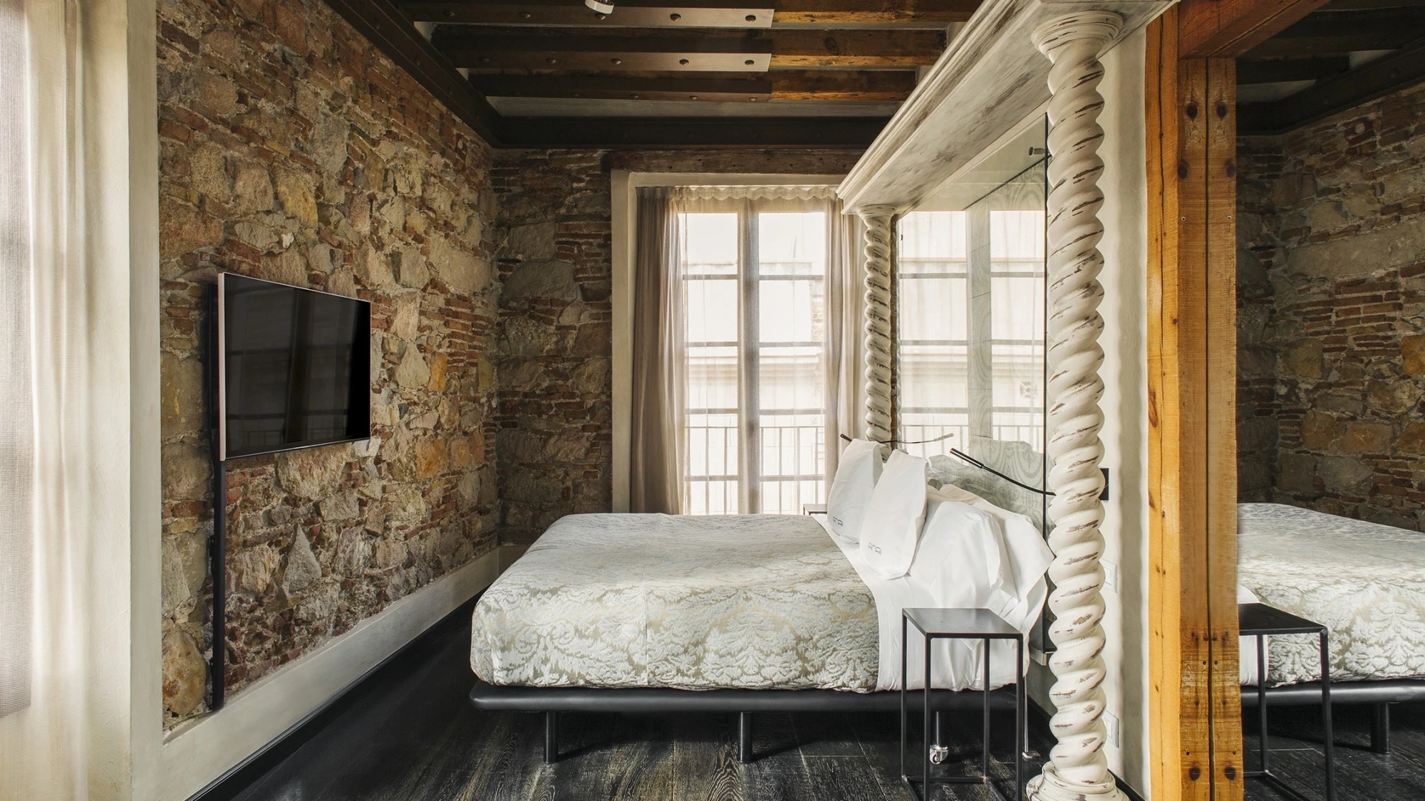 La habitación muestra una cama con ropa de cama blanca, paredes de piedra y una ventana con luz natural.