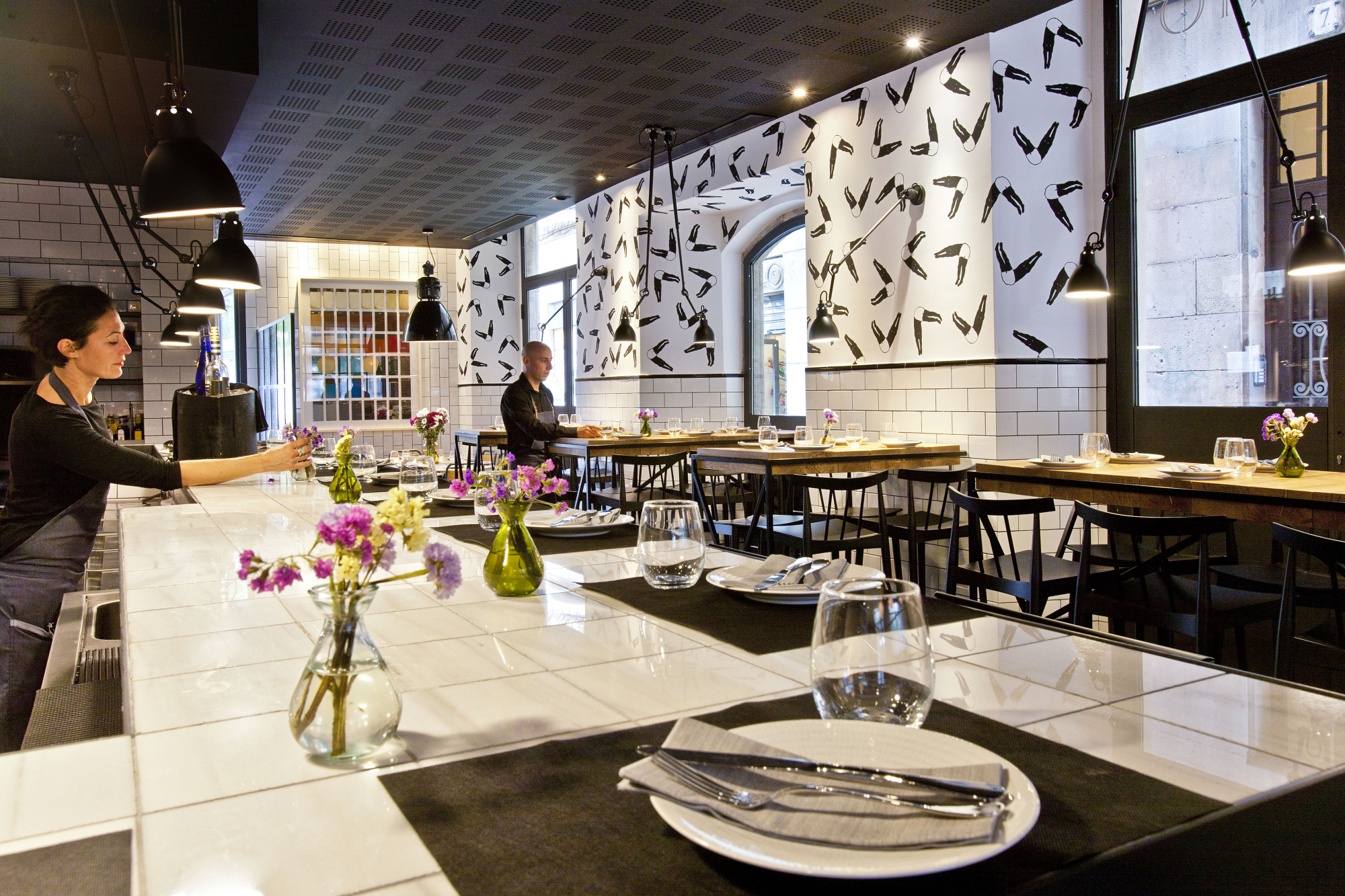Se ve un comedor con mesas cubiertas, sillas y vajilla sobre una mesa central.