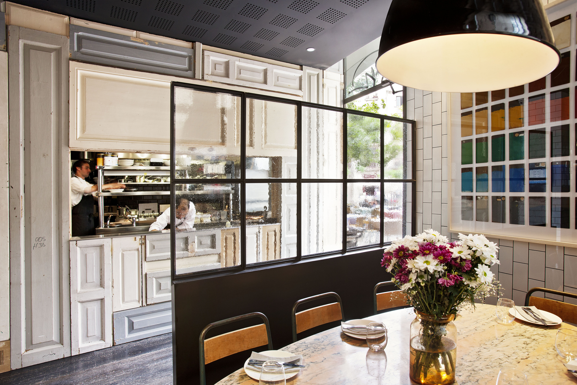 Se ve una mesa con platos y un jarrón de flores junto a una cocina con gabinetes claros y una ventana grande.