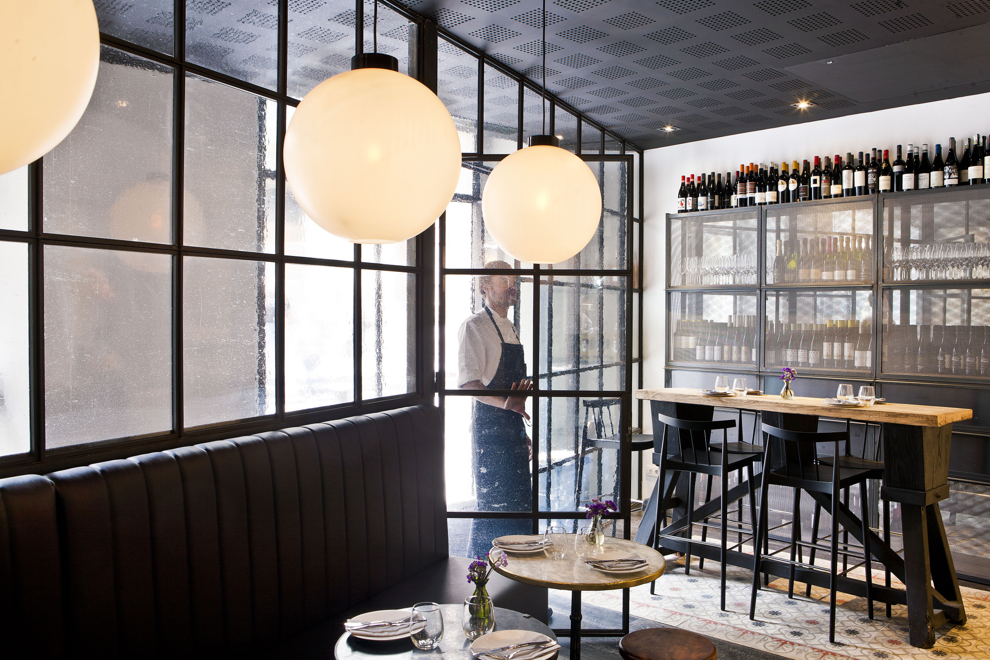 Se ve un bar con taburetes, una barra de bebidas y lámparas colgantes en un interior con ventanales.
