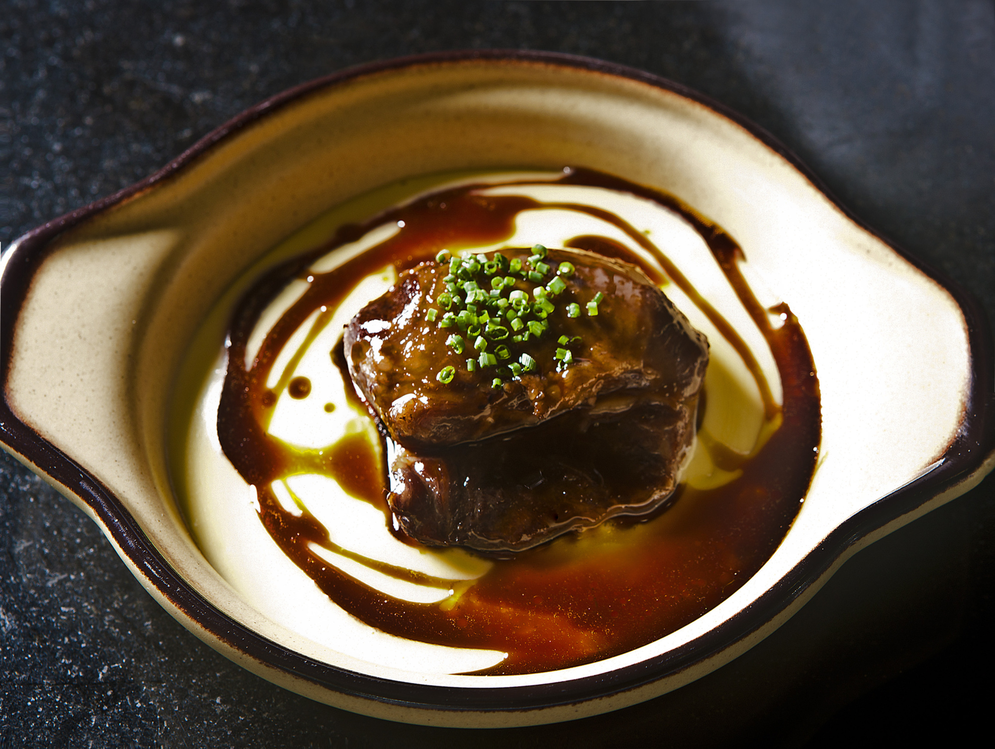 El plato presenta un trozo de carne cubierto con salsa oscura y decorado con brotes verdes.