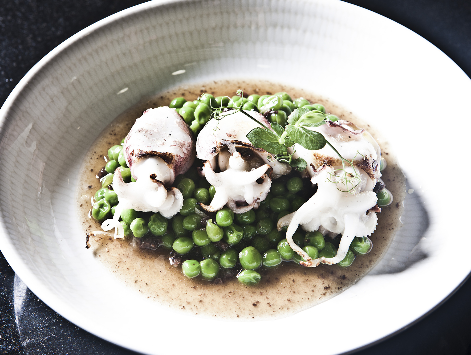 Plato blanco con sopa y pulpitos, guisantes y brotes verdes.