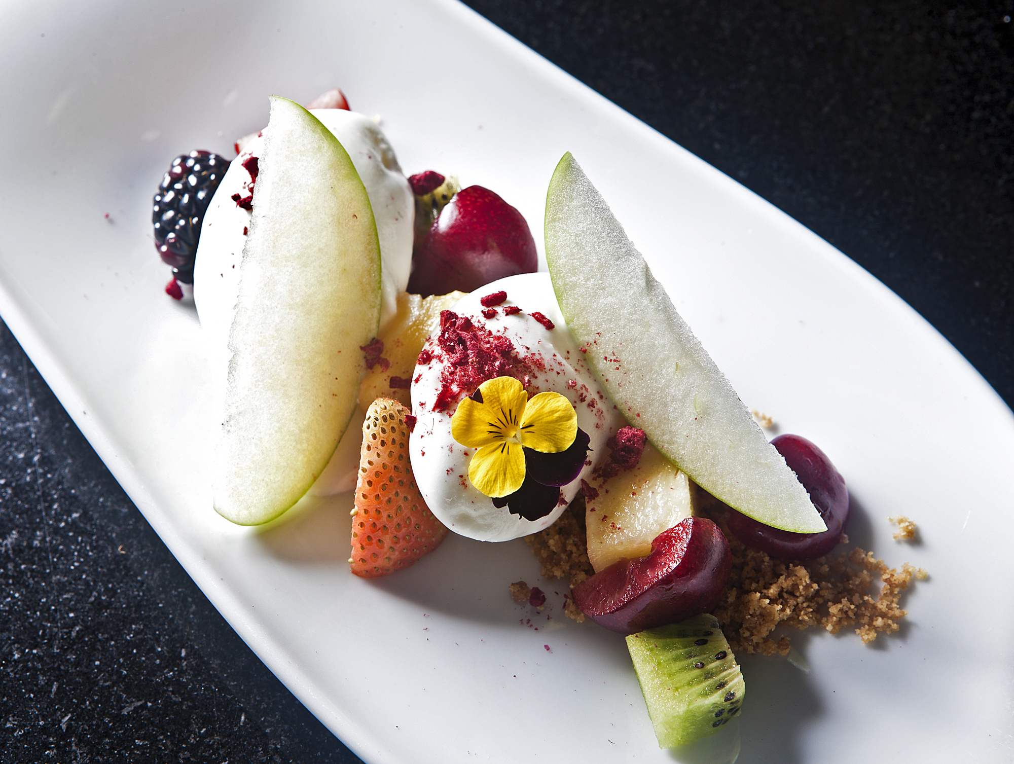Se ve un plato blanco con frutas cortadas y un postre decorado con una flor amarilla.