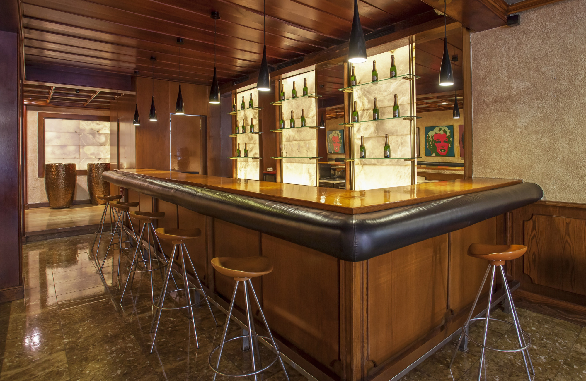 Se ve una barra de bar de madera con taburetes y botelleros iluminados al fondo.