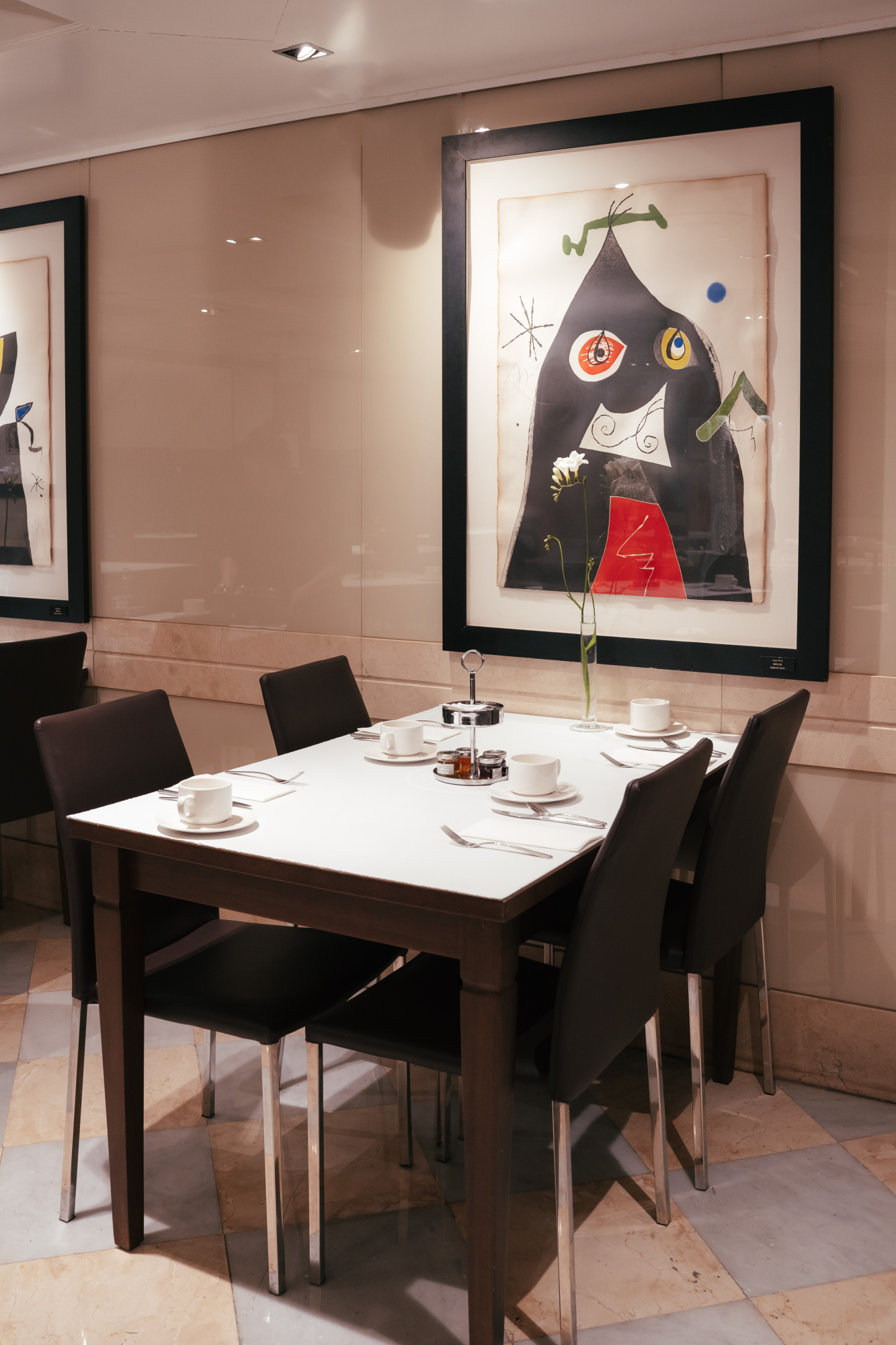 Se ve una mesa de comedor con vajilla y cuatro sillas, junto a cuadros de Joan Miró en una pared clara.