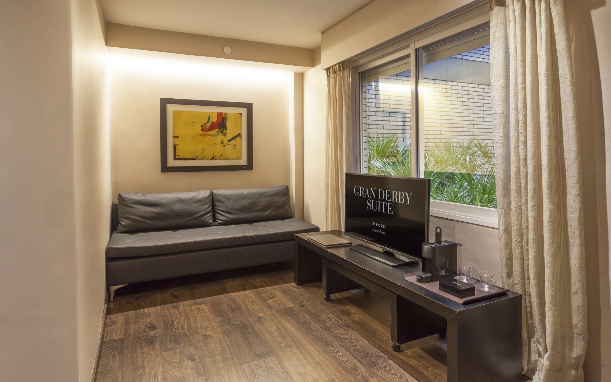 Se ve un sofá, un escritorio con silla y una ventana con cortinas en una habitación luminosa.