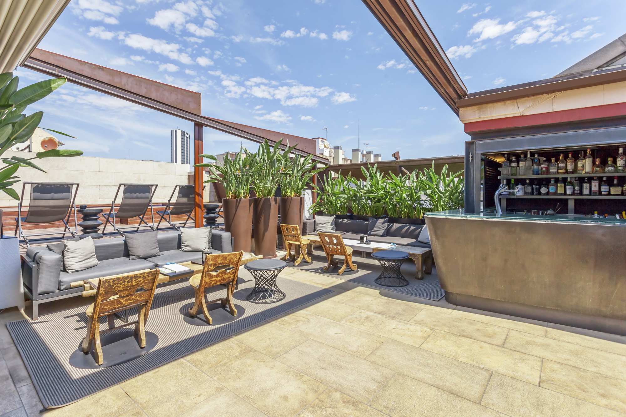 Se ve una terraza al aire libre con tumbonas, mesas y un bar con bebidas bajo un techo parcial.