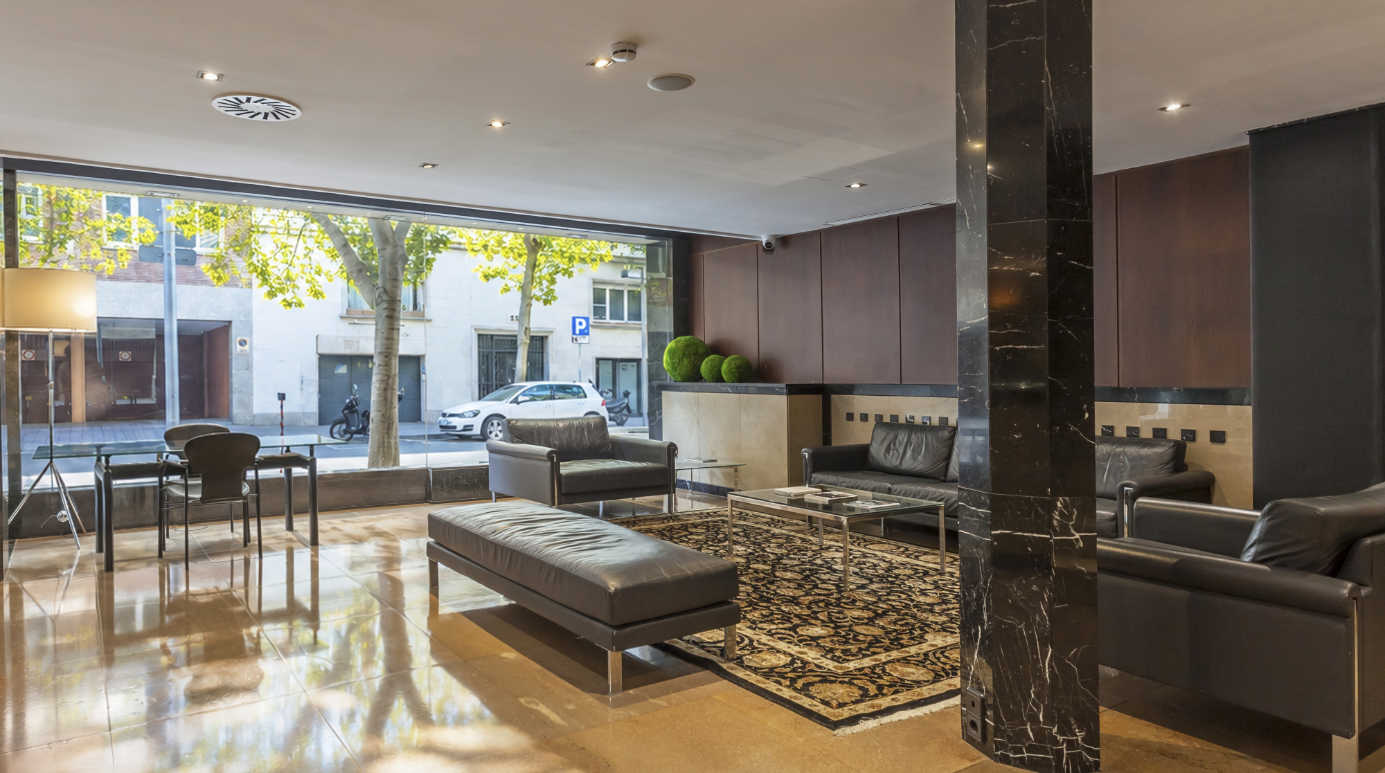 Se ve una recepción moderna con zona de estar, sillones, una mesa auxiliar y ventanales con luz natural.