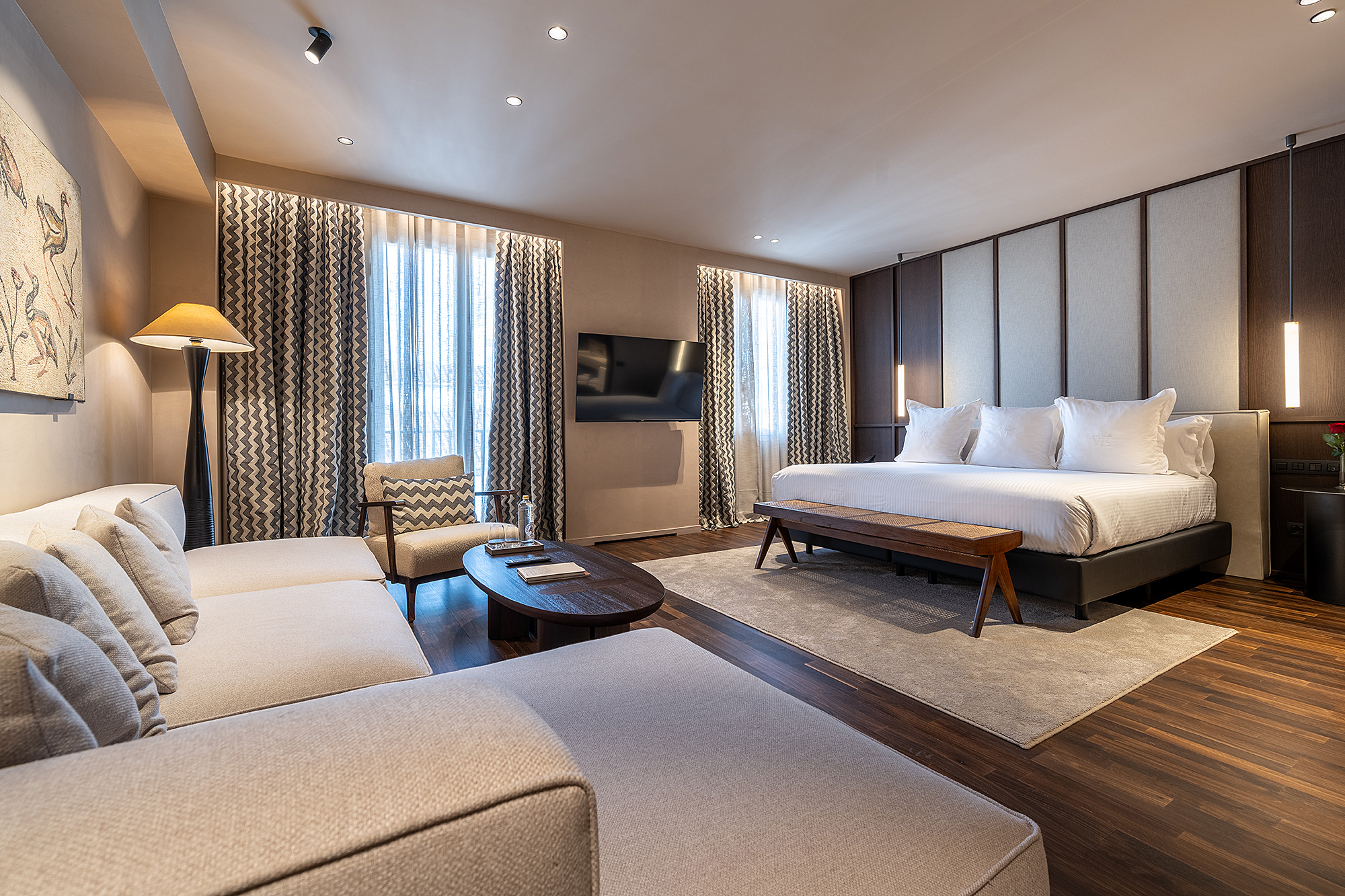 Se ve una suite con zona de estar, sofá, mesa de centro y una cama doble junto a ventanas con cortinas.