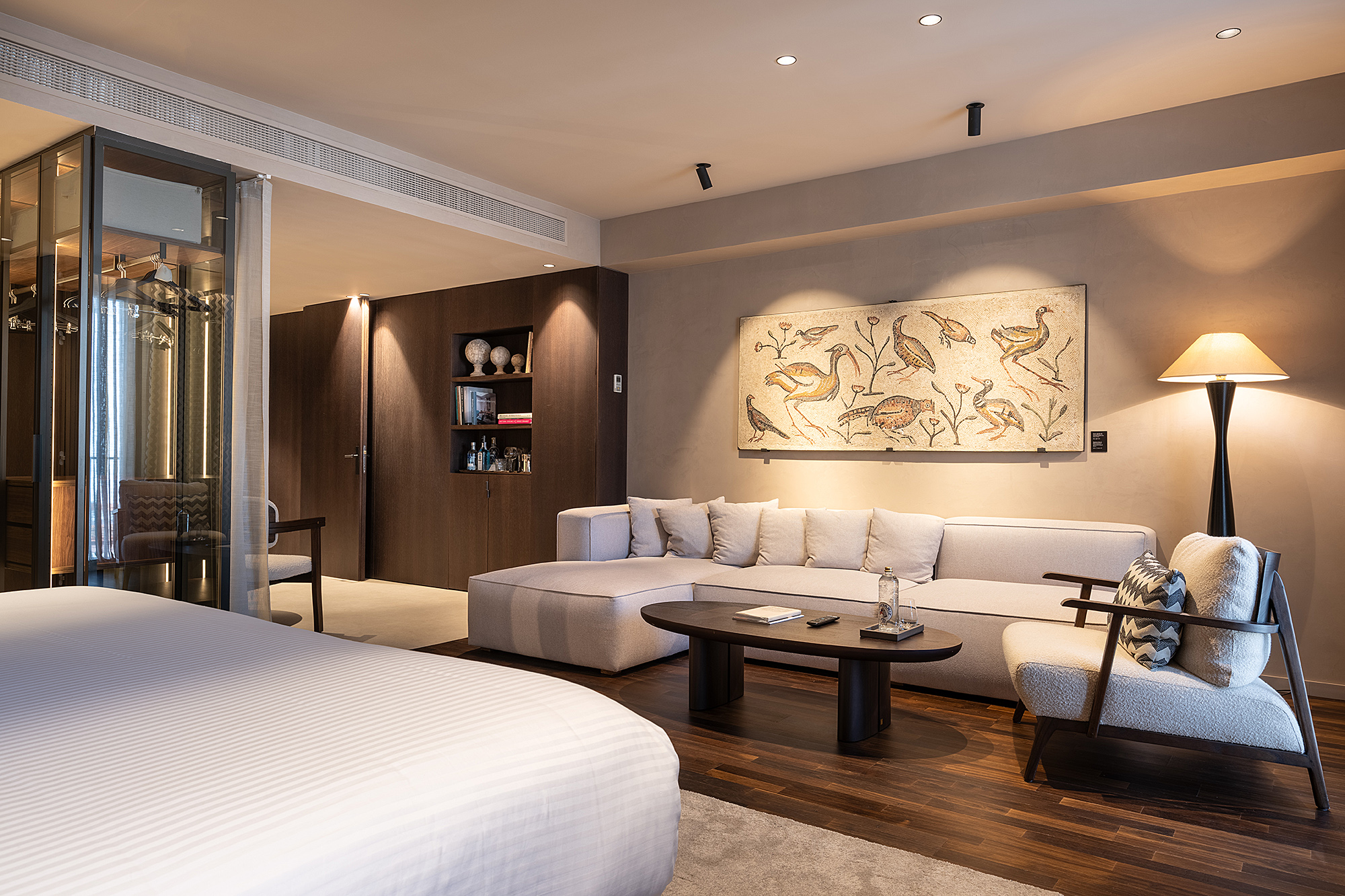 Se ve una cama doble, un sofá, una mesa de centro y una lámpara en una habitación con paredes beige y arte enmarcado.