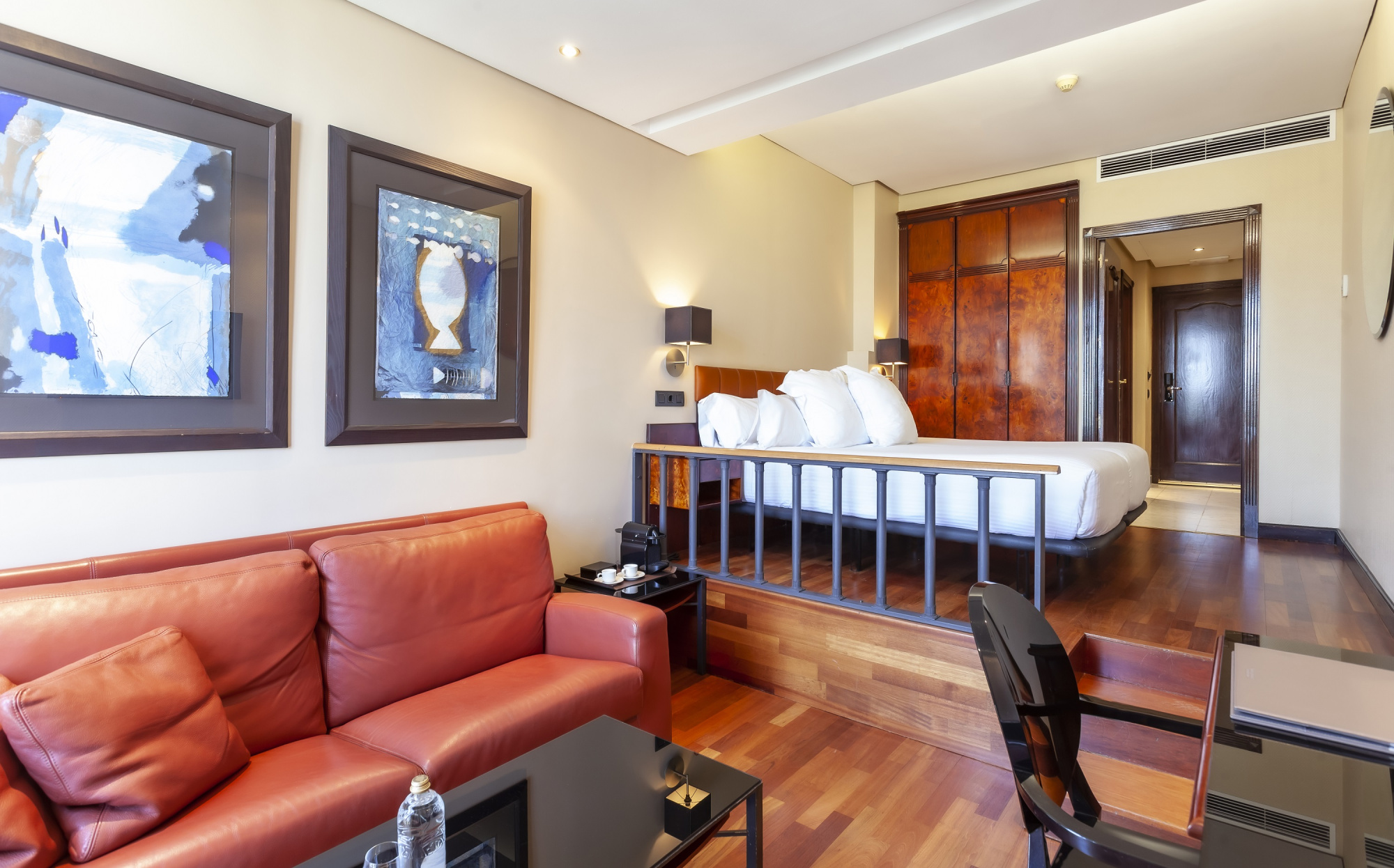 Se ve una cama doble, un sofá de cuero, una mesa auxiliar y una zona de estar junto a una ventana y puertas interiores.