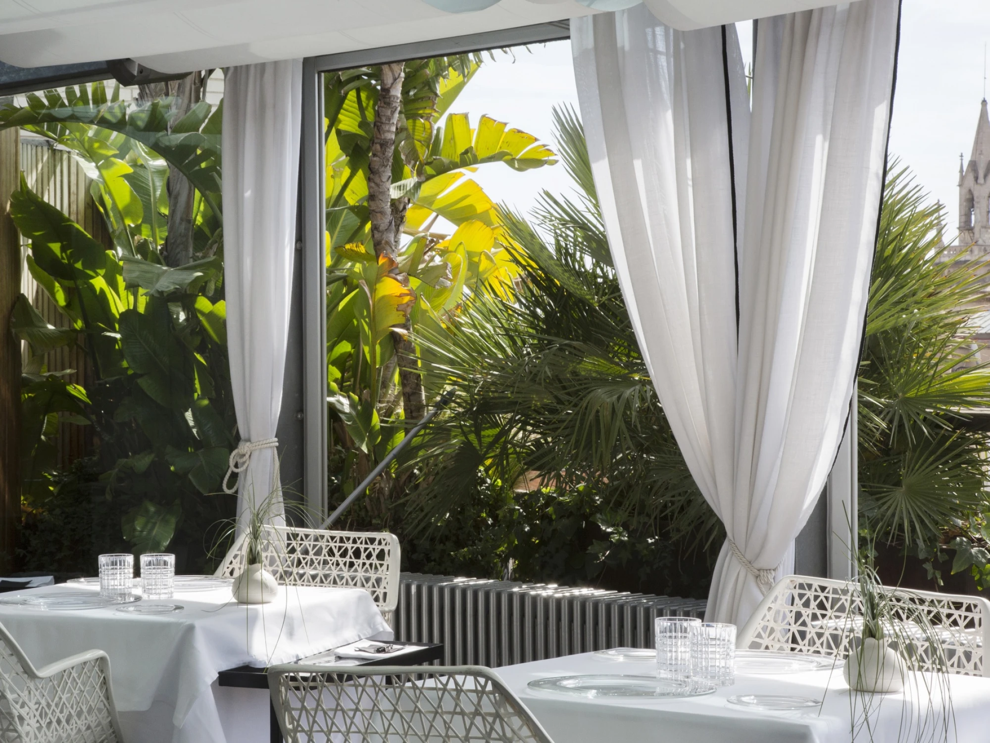 Una terraza cubierta con mesas blancas, sillas de rejilla y cortinas traslúcidas sobre vegetación tropical.