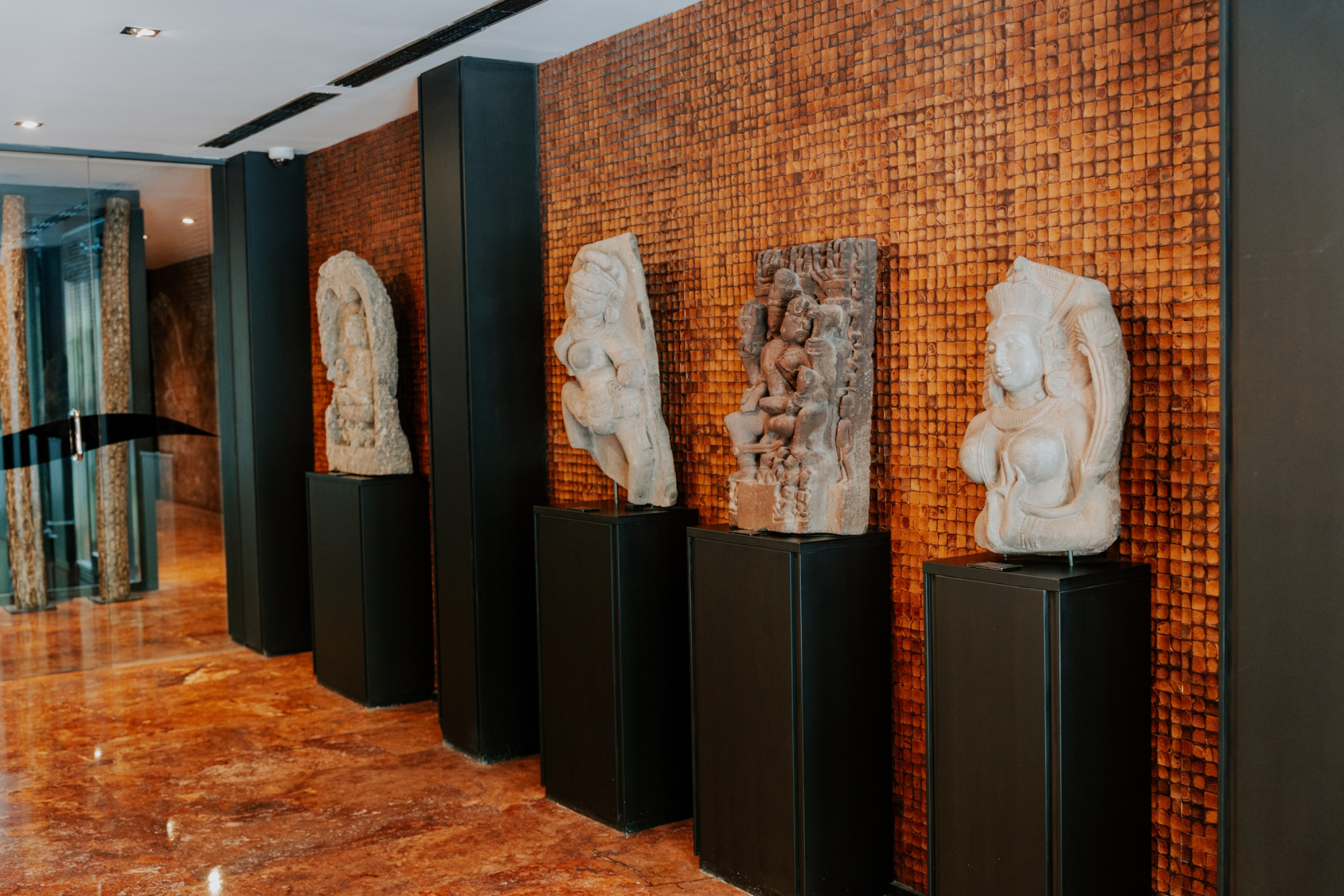 Se ven varias esculturas sobre pedestales negros a lo largo de un pasillo con suelo de madera y pared de mosaico.