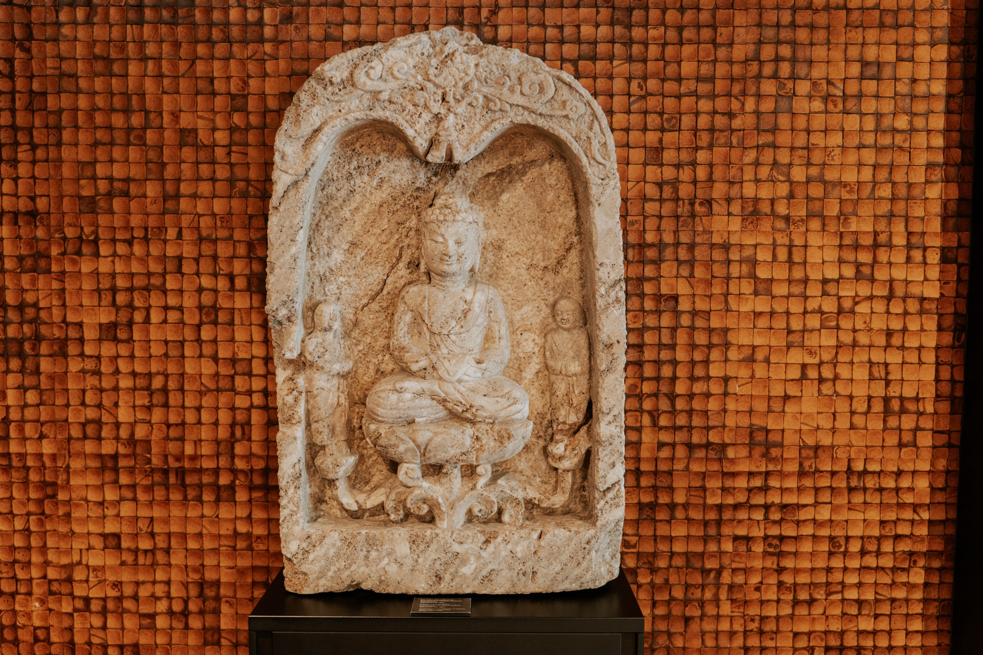 Se ve una escultura tallada sobre un fondo de pequeñas baldosas naranjas, con una placa de exhibición negra.