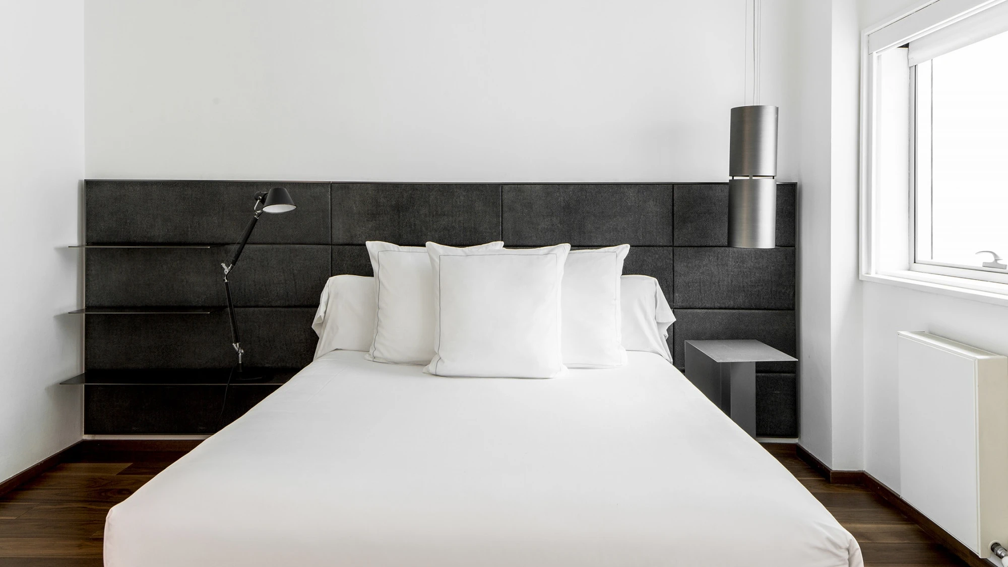 Un dormitorio moderno muestra una cama doble con ropa de cama blanca y dos almohadas, junto a una pared gris y una ventana con cortina ligera.