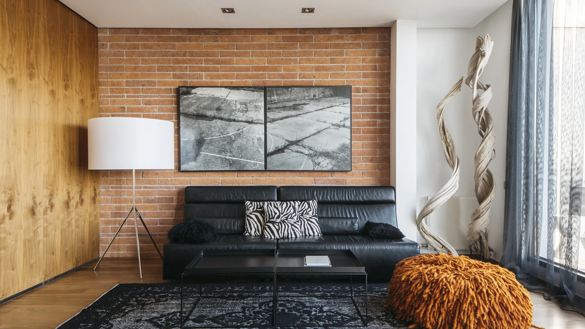 Un salón moderno muestra un sofá de cuero negro, una lámpara de pie, cuadros decorativos y una pared de ladrillo visto.