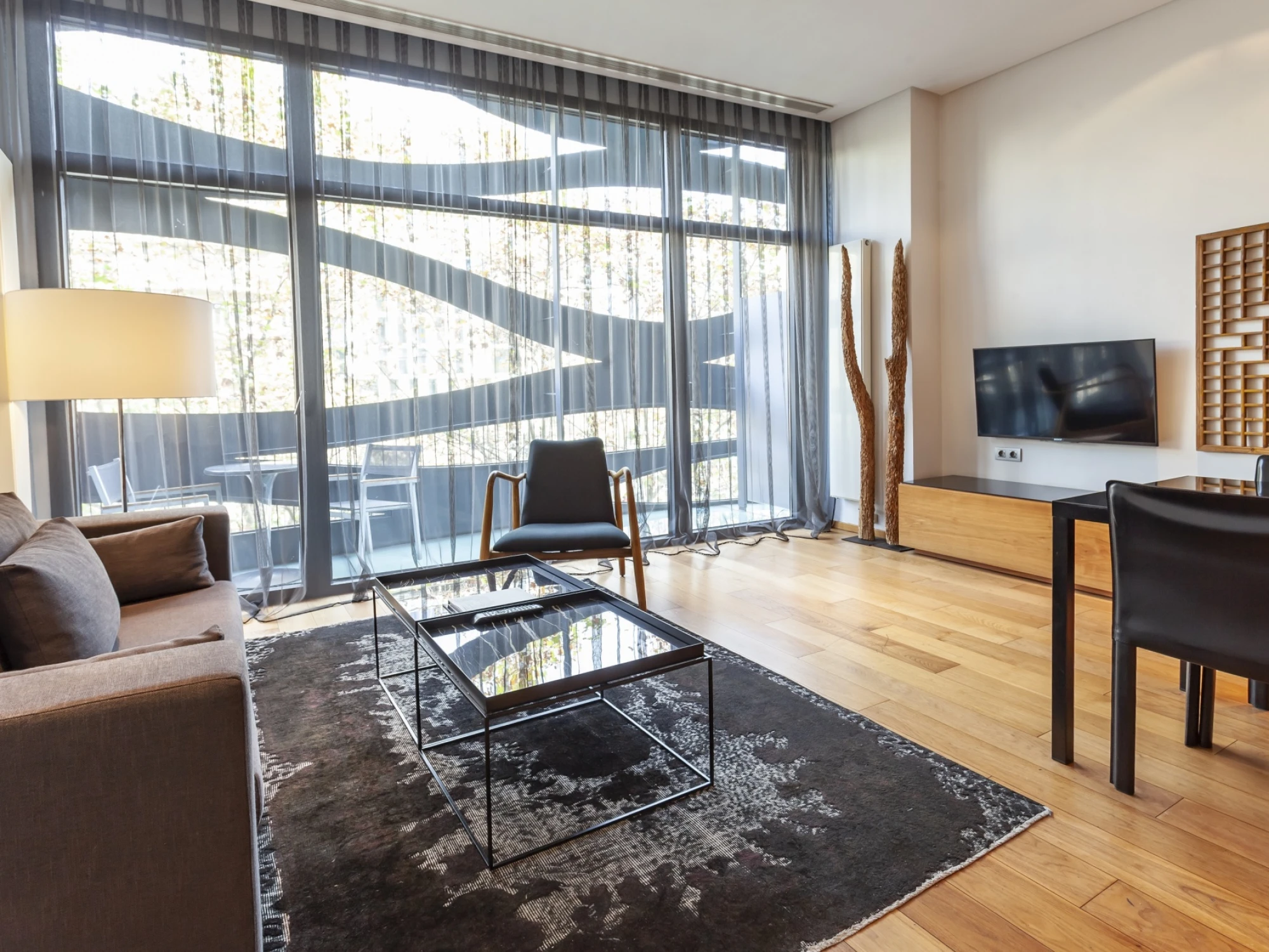 La sala de estar muestra grandes ventanales, muebles modernos, una alfombra y una mesa de centro junto a un televisor.