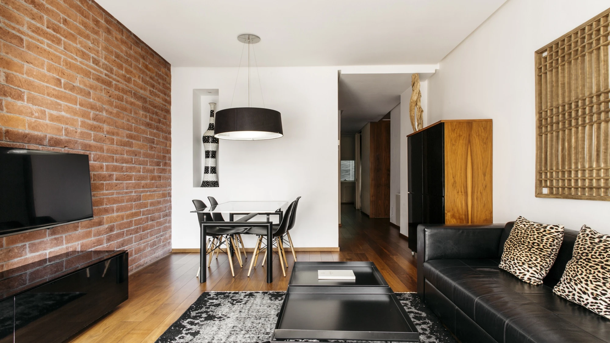 La imagen muestra una sala de estar con sofá negro, alfombra, mesa de centro, televisión y chimenea decorativa de ladrillo.