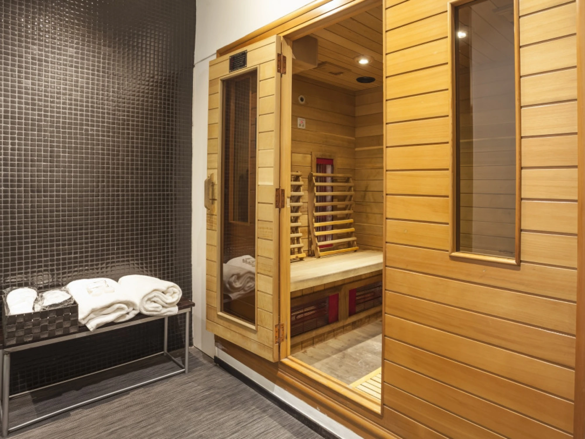 Se ve una sauna de madera con bancadas y una zona contigua revestida, con toallas dobladas sobre un banco metálico.