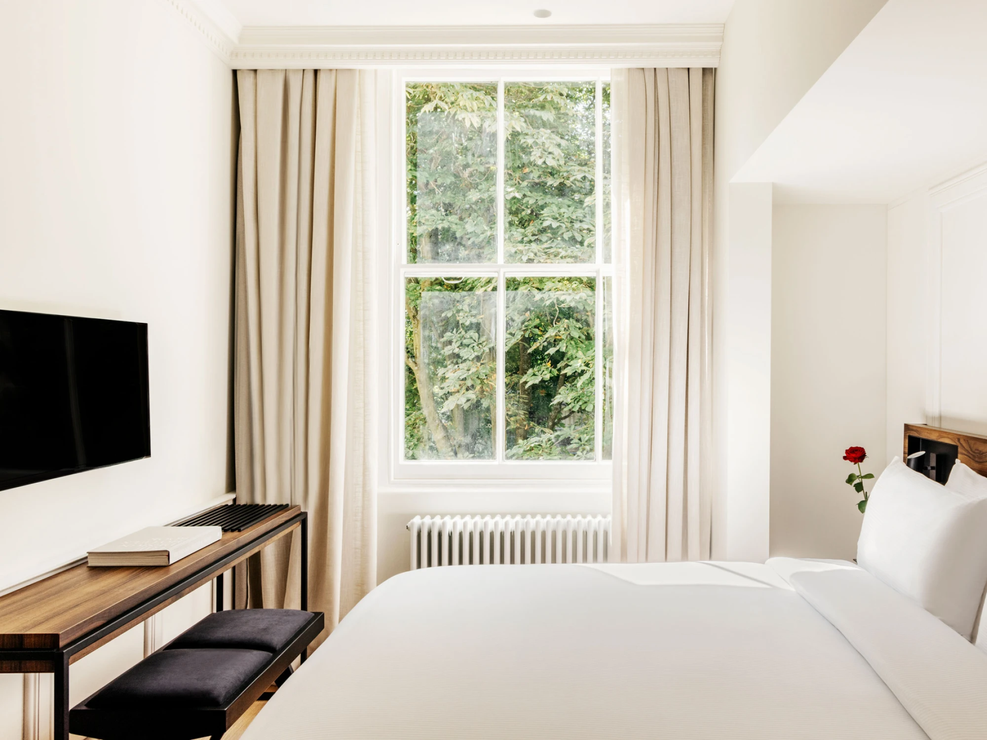 Se ve una cama doble con ropa de cama blanca, una ventana con cortinas y un televisor montado en la pared.