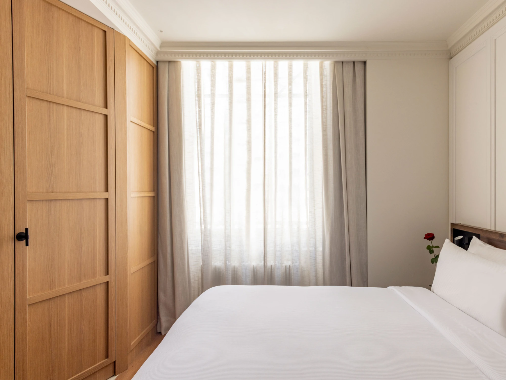 Se ve una cama doble con ropa de cama blanca, una cortina y una puerta de madera en una habitación luminosa.