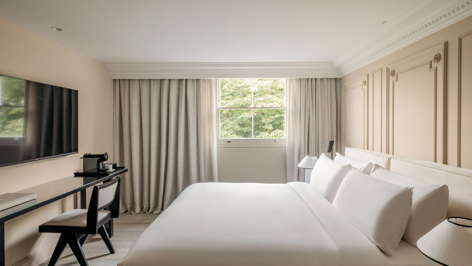 Se ve una cama doble con almohadas, una ventana con cortinas y un escritorio con silla junto a una pared con TV.