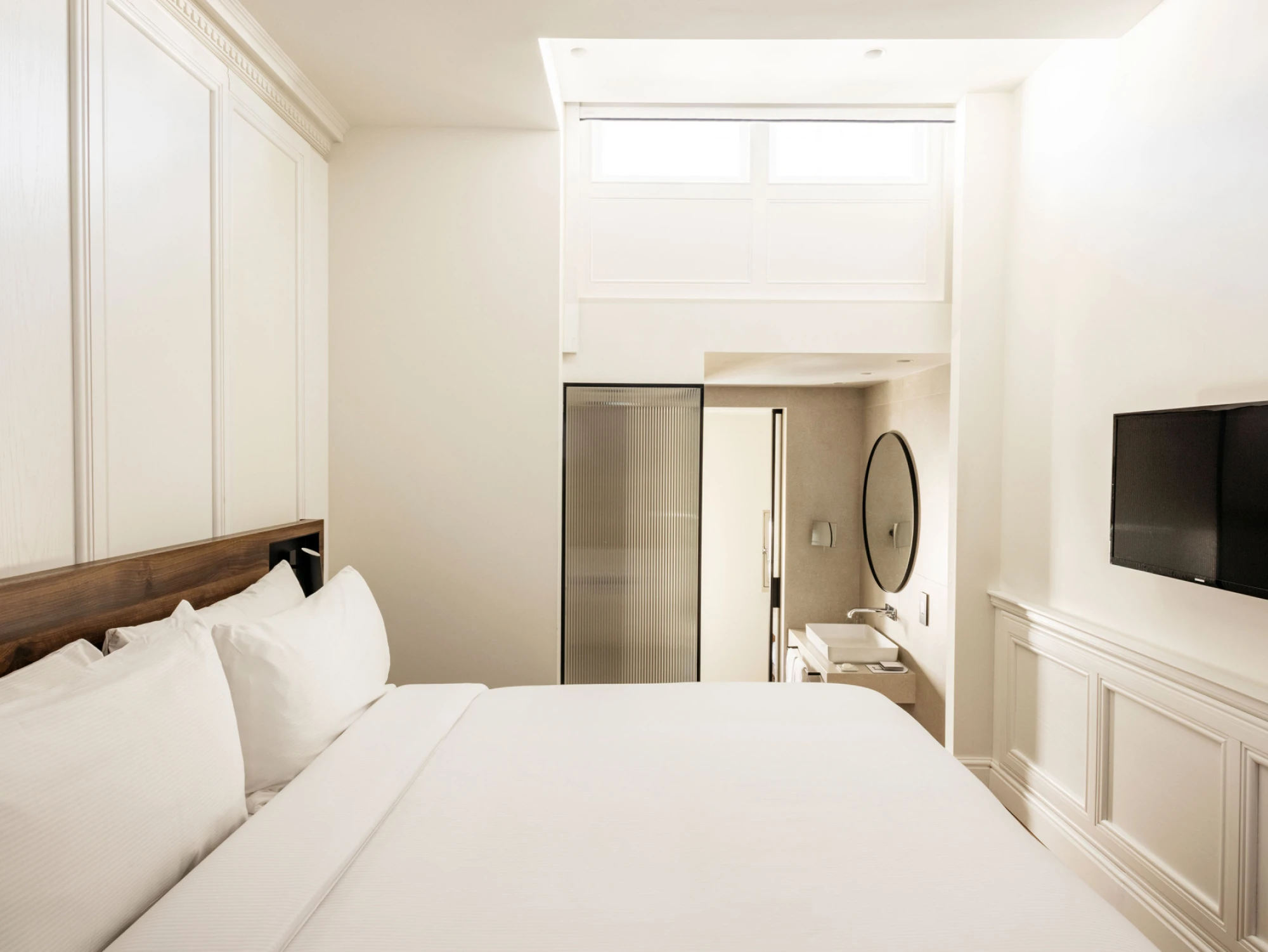 Se ve una cama doble con ropa de cama blanca y un televisor de pared en una habitación luminosa.