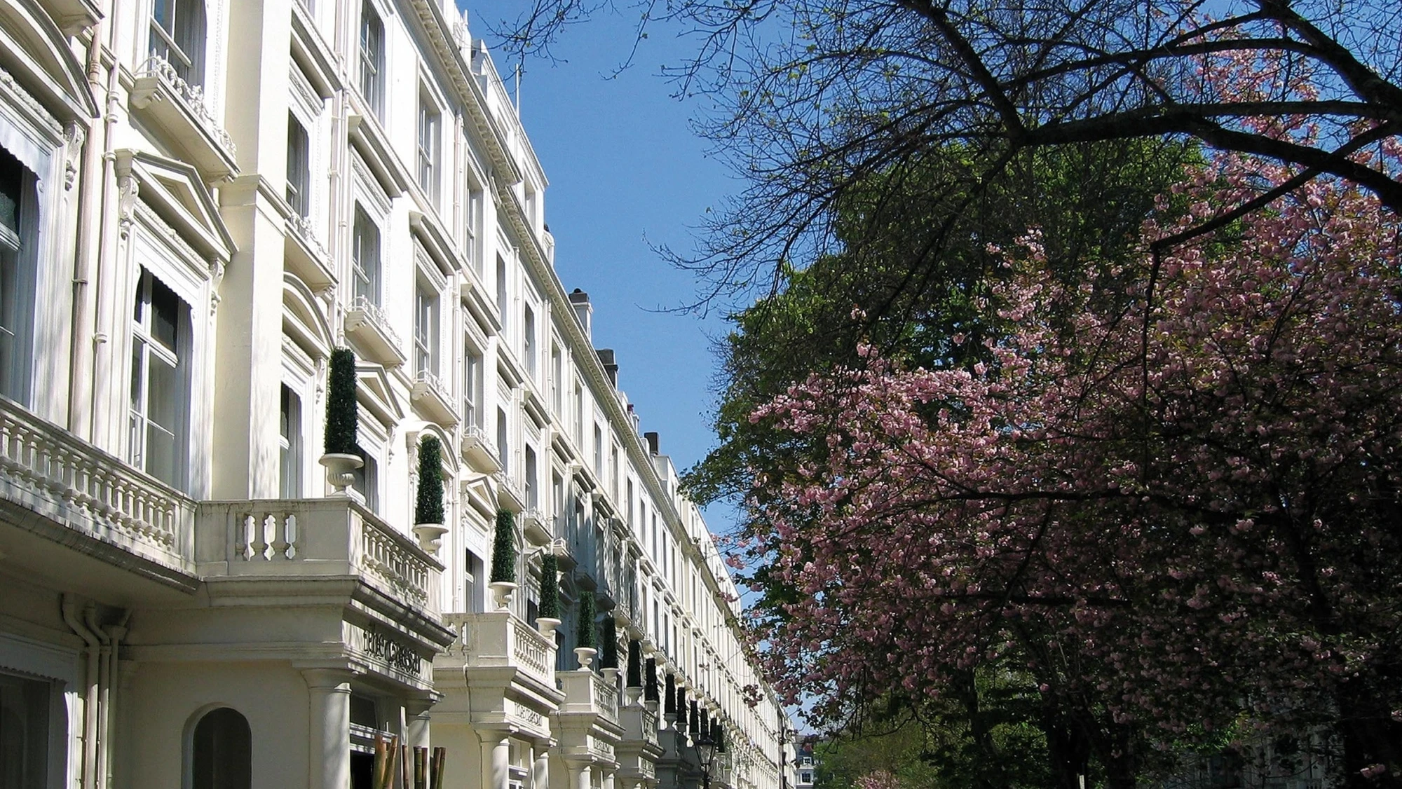 Vista exterior de una fachada de estilo victoriano con balcones y ventanas, junto a árboles en flor.