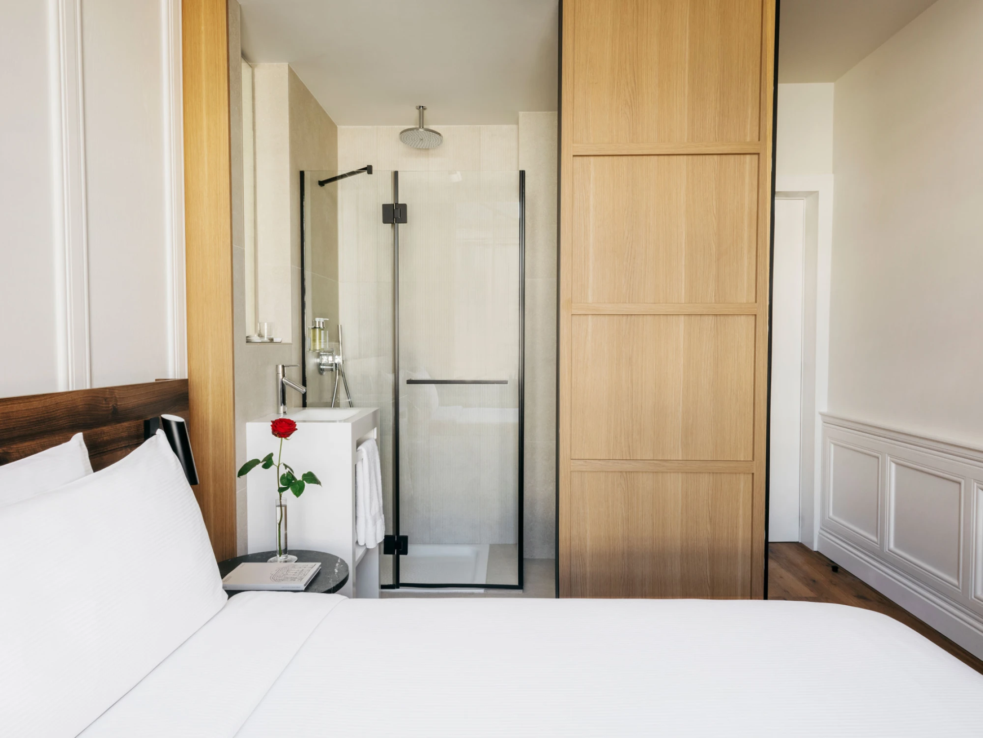 Se ve una cama con ropa de cama blanca, una puerta de madera y una mampara de ducha de vidrio en el interior.