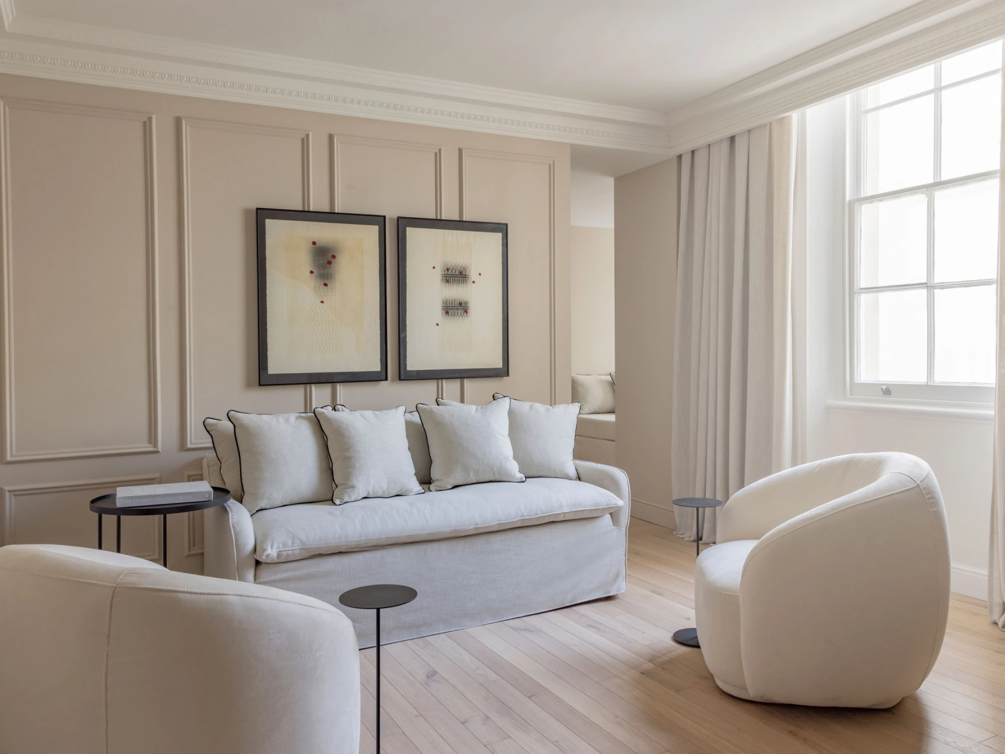 Sala de estar luminosa con sofá blanco, sillón y cuadros en la pared junto a una ventana grande.